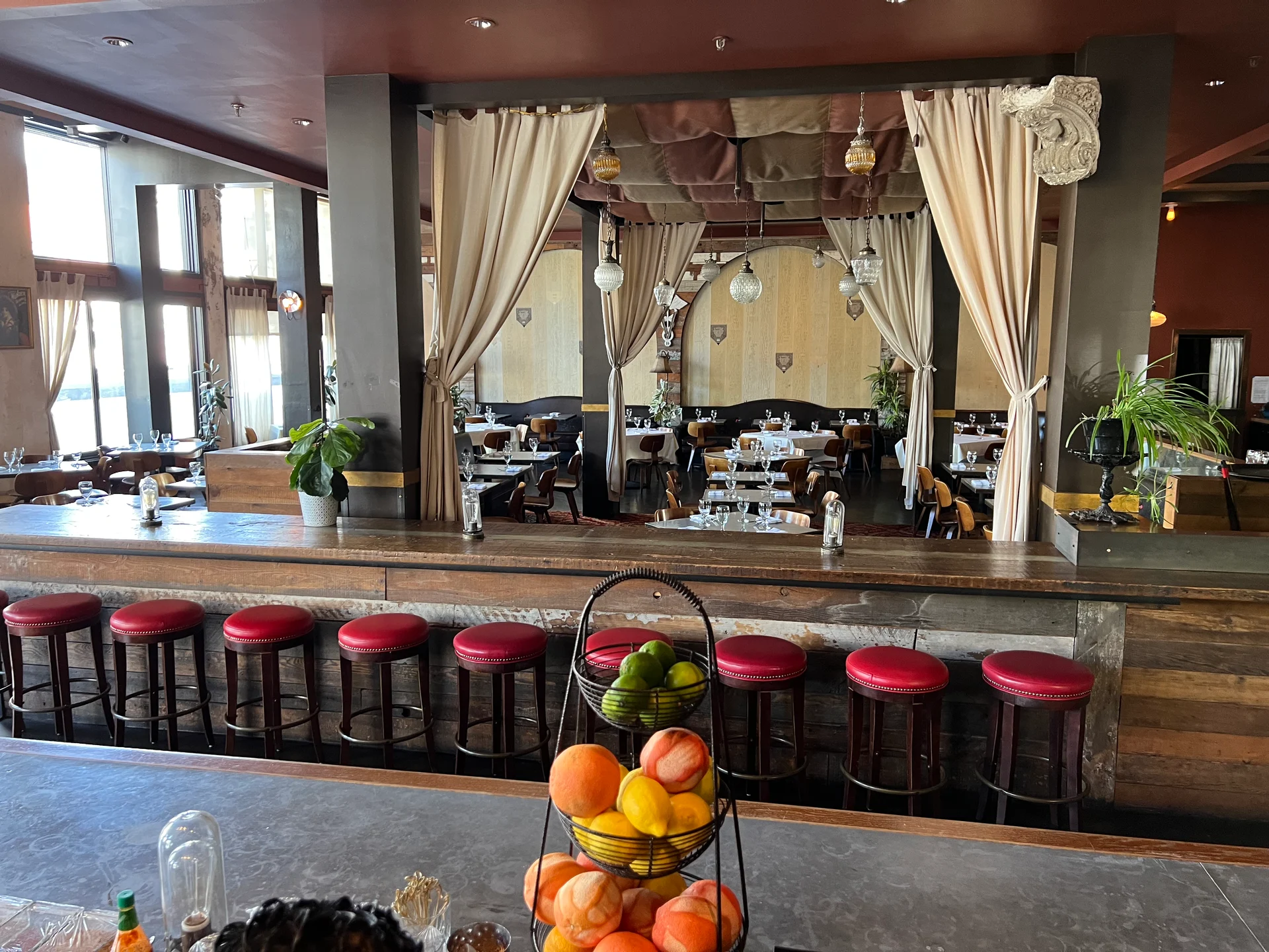 Spacious restaurant with red stools at a bar, fruit basket in the foreground, tables and chairs in the background, and elegant draped curtains.