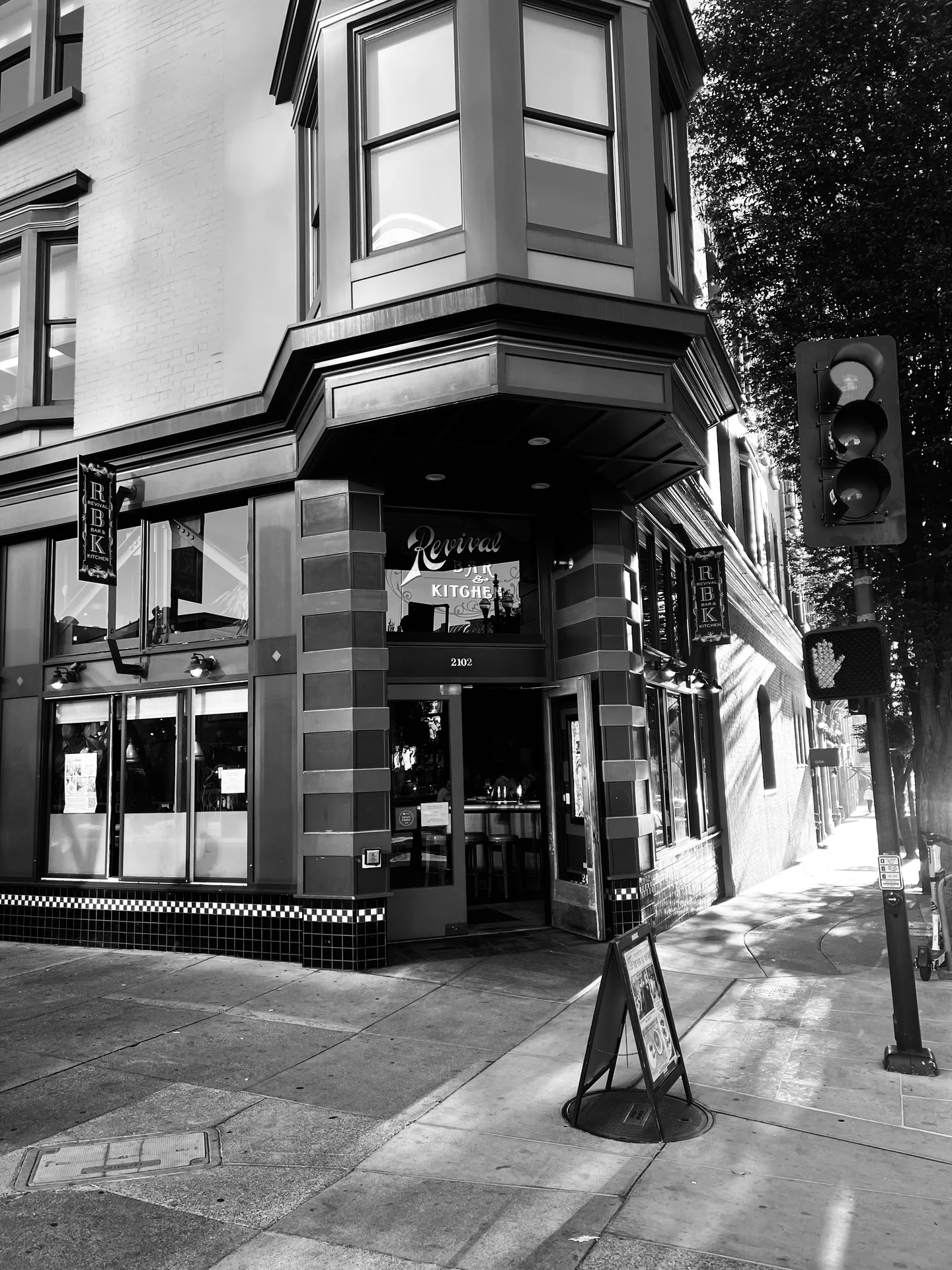 A black-and-white photo of a corner restaurant named "Revival Kitchen." It features large windows, a hanging sign, and a sidewalk sandwich board. A traffic light and crosswalk signal are on the right.