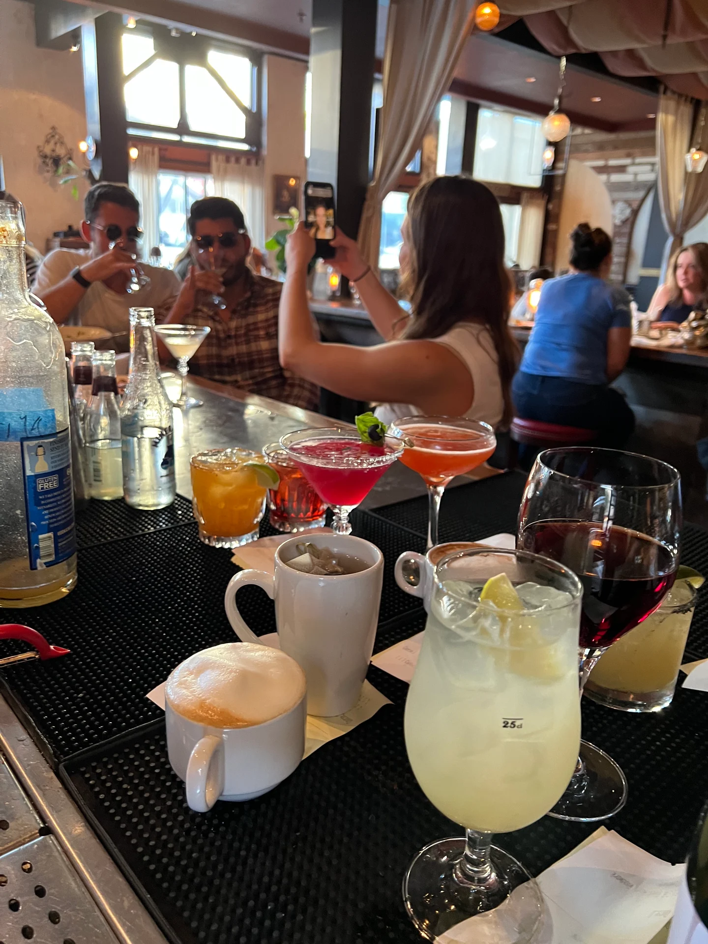 A group of people sitting at a bar, enjoying drinks such as cocktails and fruit juices. They are seated around a table with various drinkware and food items in an indoor restaurant setting.