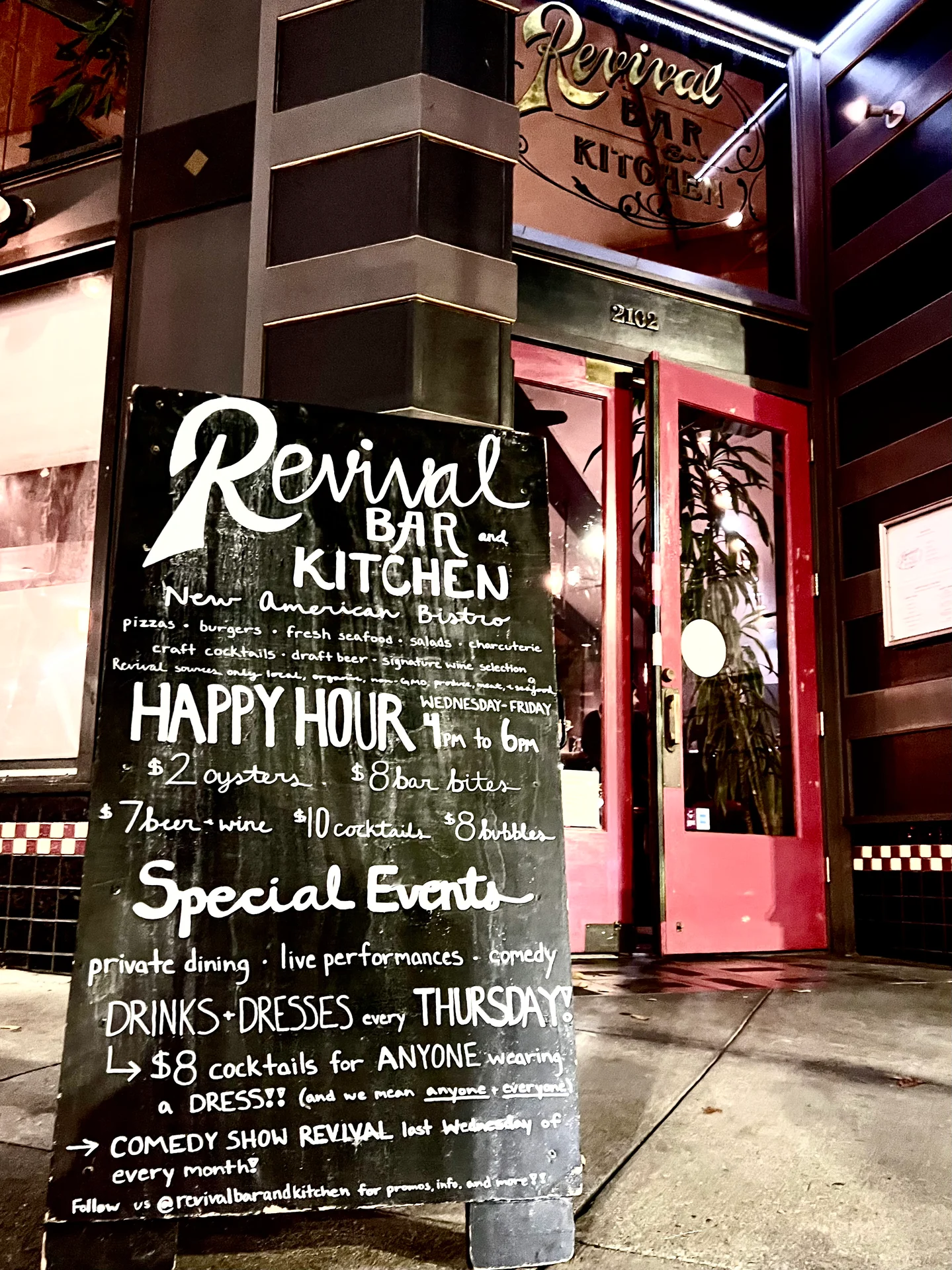 A chalkboard sign outside a restaurant displaying various food and drink specials including $2 oysters, $8 bar bites, and information about happy hour and special events. The sign is titled "Revival BAR KITCHEN" and lists menu items like pizzas and burgers. It also promotes comedy shows and themed drink nights for attendees wearing dresses.