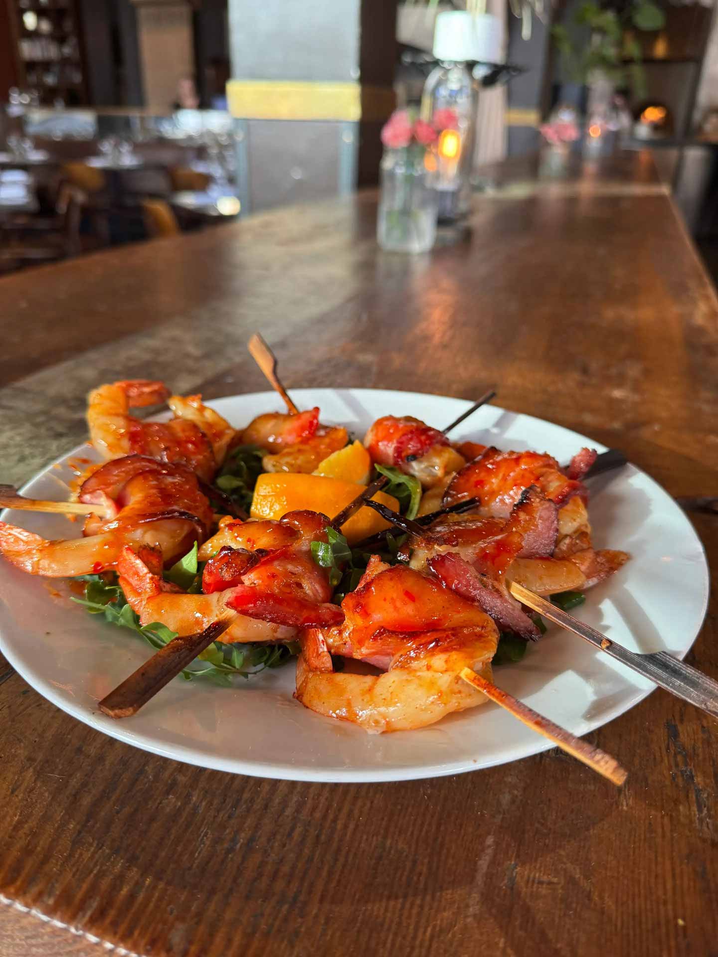 A white plate on a wooden table holds skewers of glazed shrimp wrapped in bacon, garnished with greens and orange slices. The setting suggests a cozy restaurant ambiance.