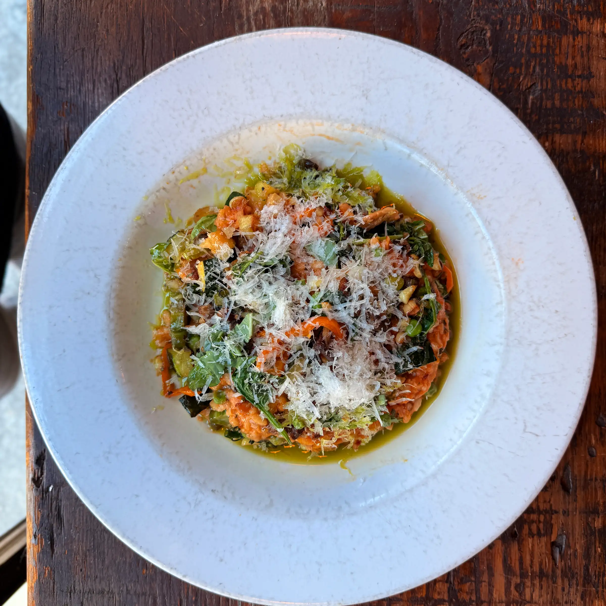 A white plate on a wooden table holds a vibrant vegetable risotto topped with grated cheese and herbs, evoking a warm and rustic dining ambiance.