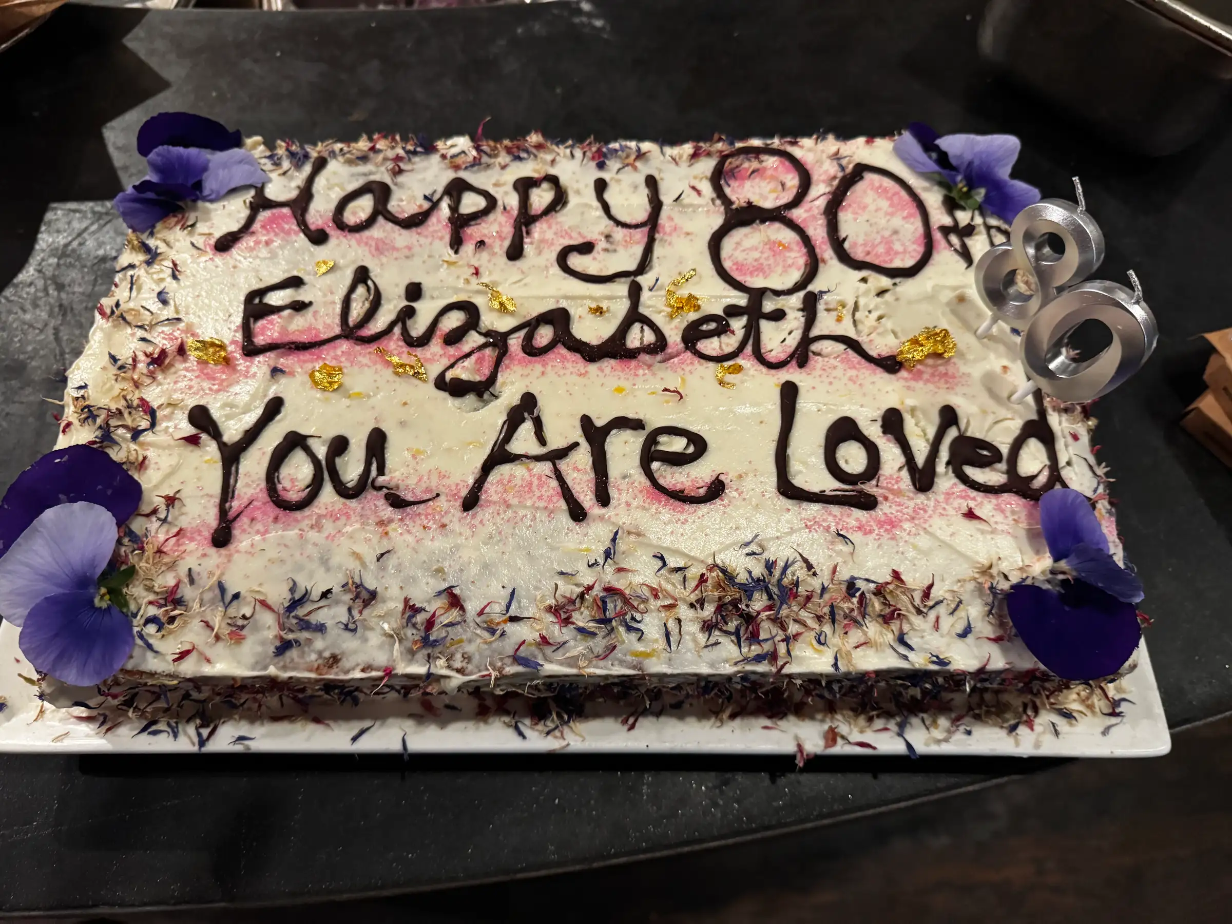 Rectangular cake celebrating an 80th birthday for "Elizabeth." Decorated with purple flowers, candles, and the message "You Are Loved." Festive and joyful.