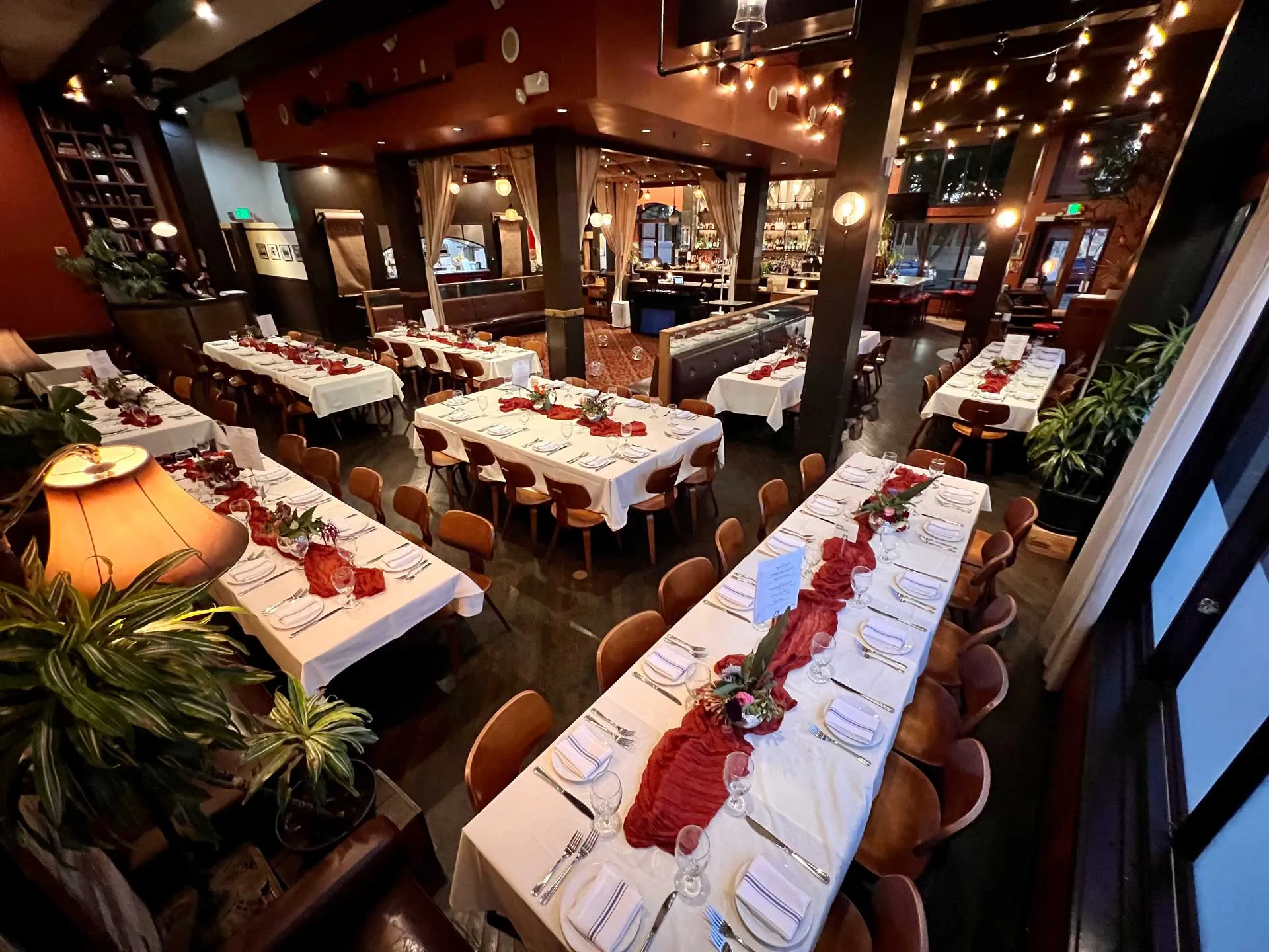 Elegant restaurant set for a formal event with long tables covered in white cloths, neatly arranged chairs, and red centerpieces. Warm ambient lighting and plants create a cozy atmosphere.