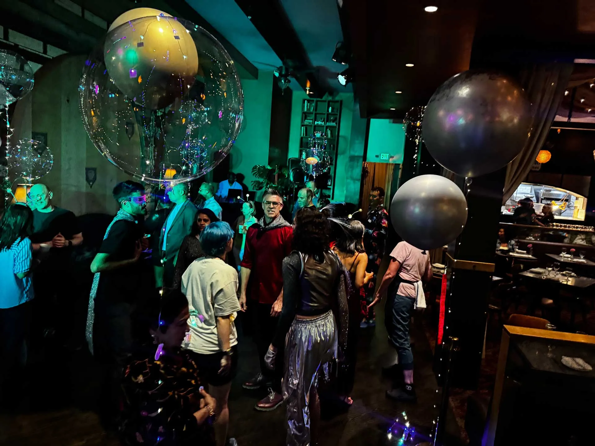 A lively party scene with people dancing under colorful lights. Large balloons float above, creating a festive atmosphere. Guests appear joyful and engaged.
