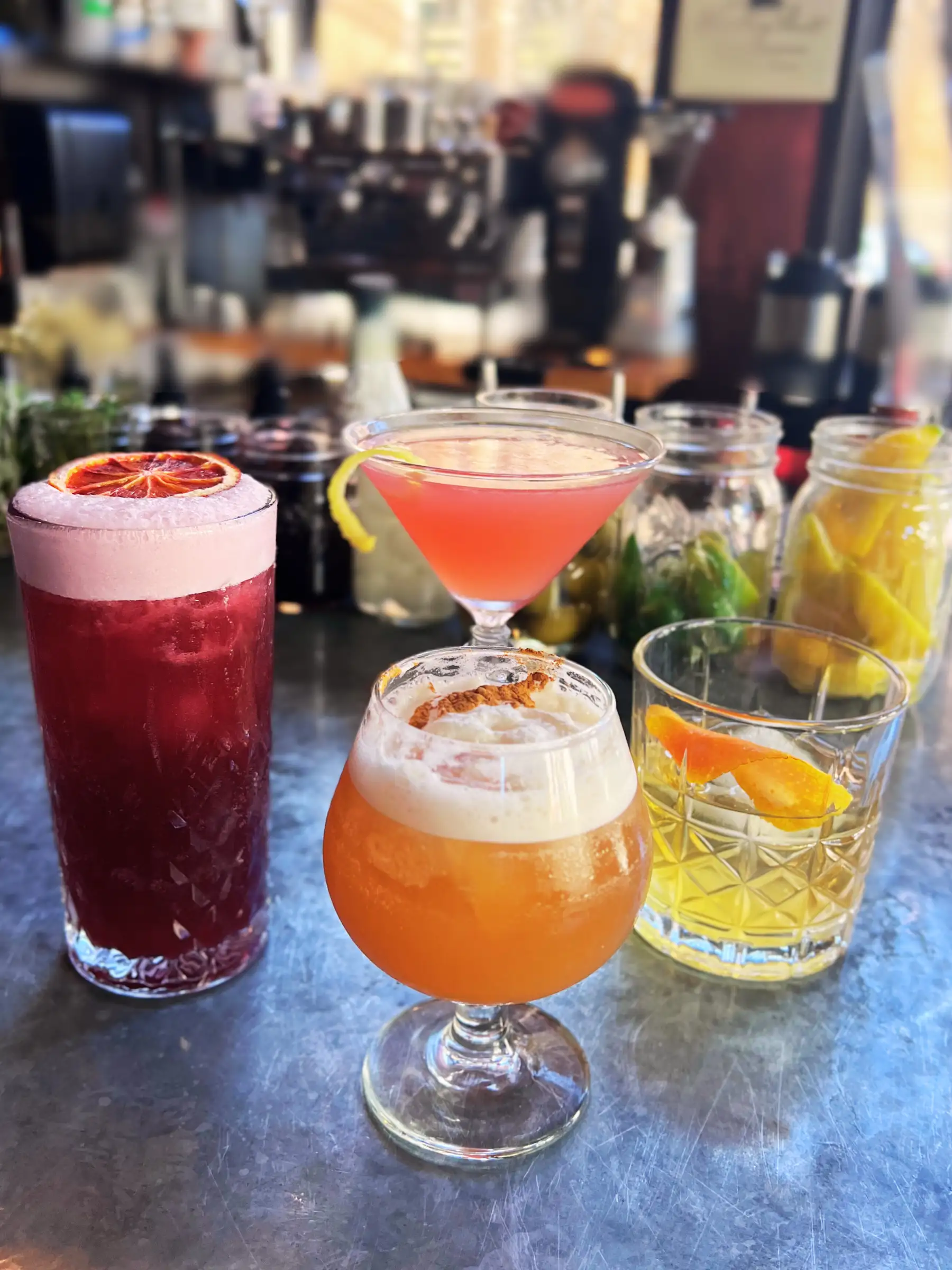 A variety of mixed drinks on a bar counter: a pink cocktail in a martini glass, a foam-topped amber drink, a red cocktail, and a yellow drink with an orange twist.
