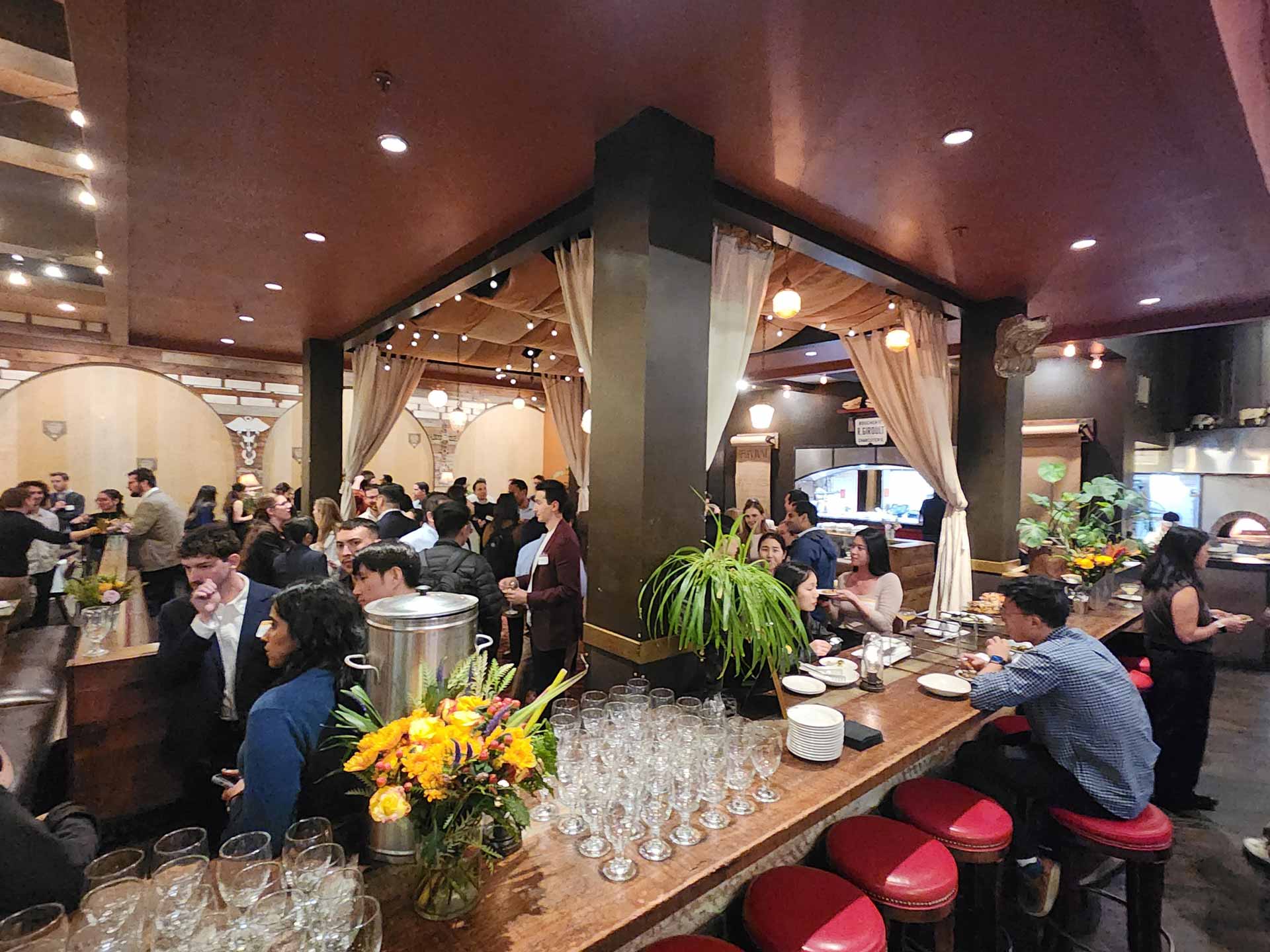 A lively bar scene with people socializing around a central pillar. Glassware, floral arrangements, and soft lighting create a warm, inviting atmosphere.