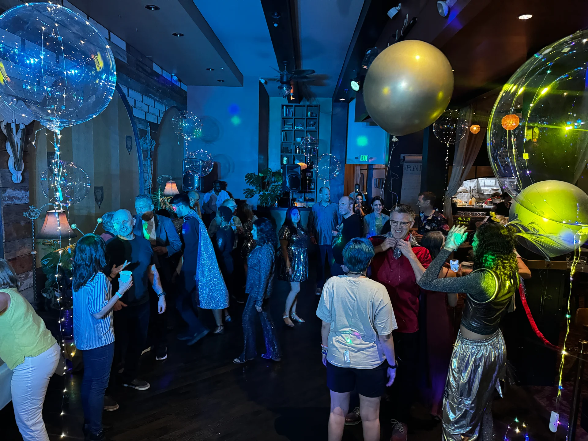A lively party scene with people dancing under colorful lights. Large balloons float above, creating a festive atmosphere. Guests appear joyful and engaged.