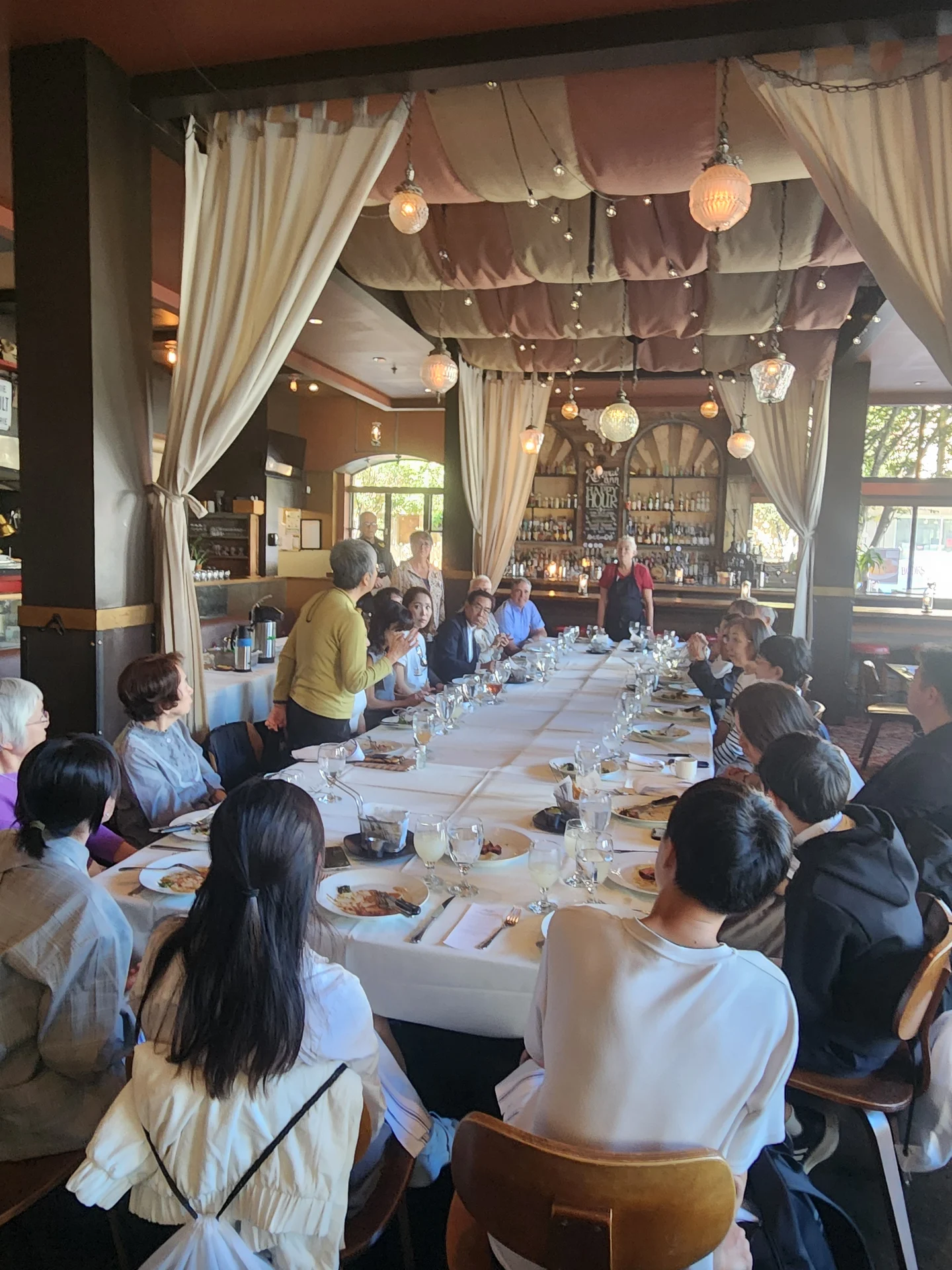 Gathering in an elegant restaurant with draped ceiling and soft lighting, a long table hosts a diverse group engaged in lively conversation and dining.