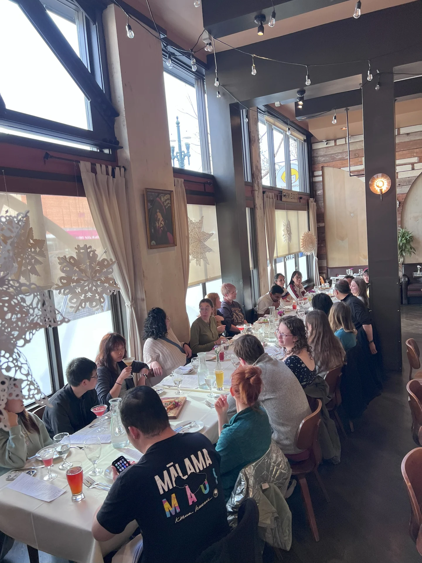 A large group of people sit around a long table in a cozy restaurant, chatting and enjoying drinks. The atmosphere is lively and warm, with soft lighting and snowflake decorations on the windows.