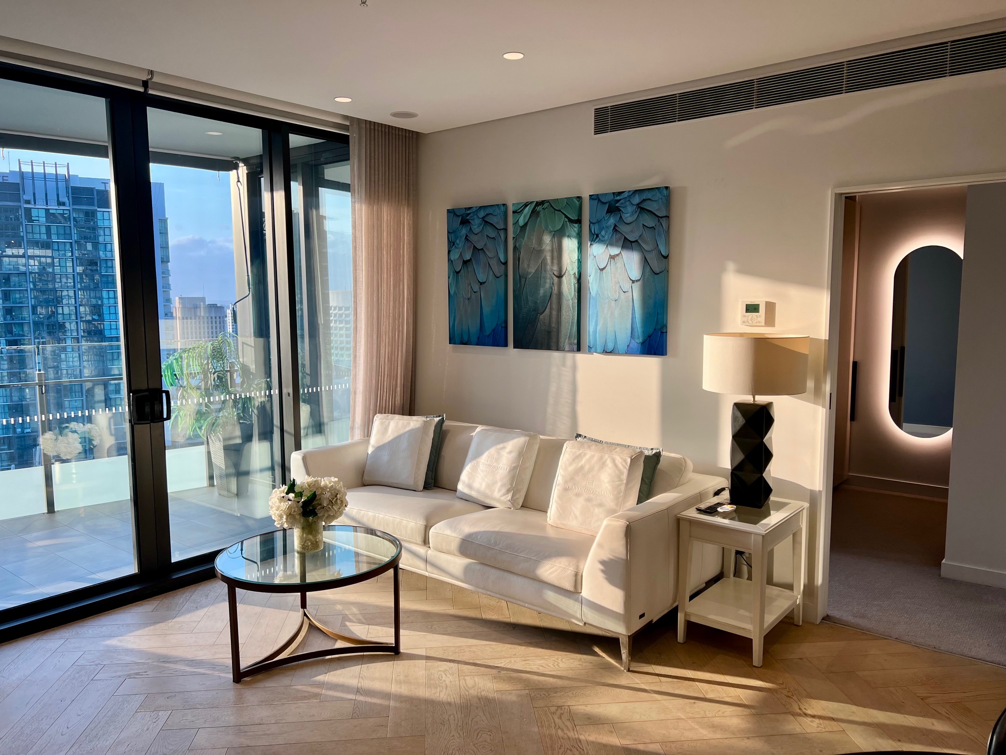 Modern high-rise apartment living room with white leather couch, glass coffee table, blue feather wall artwork, and floor-to-ceiling glass doors opening to a balcony