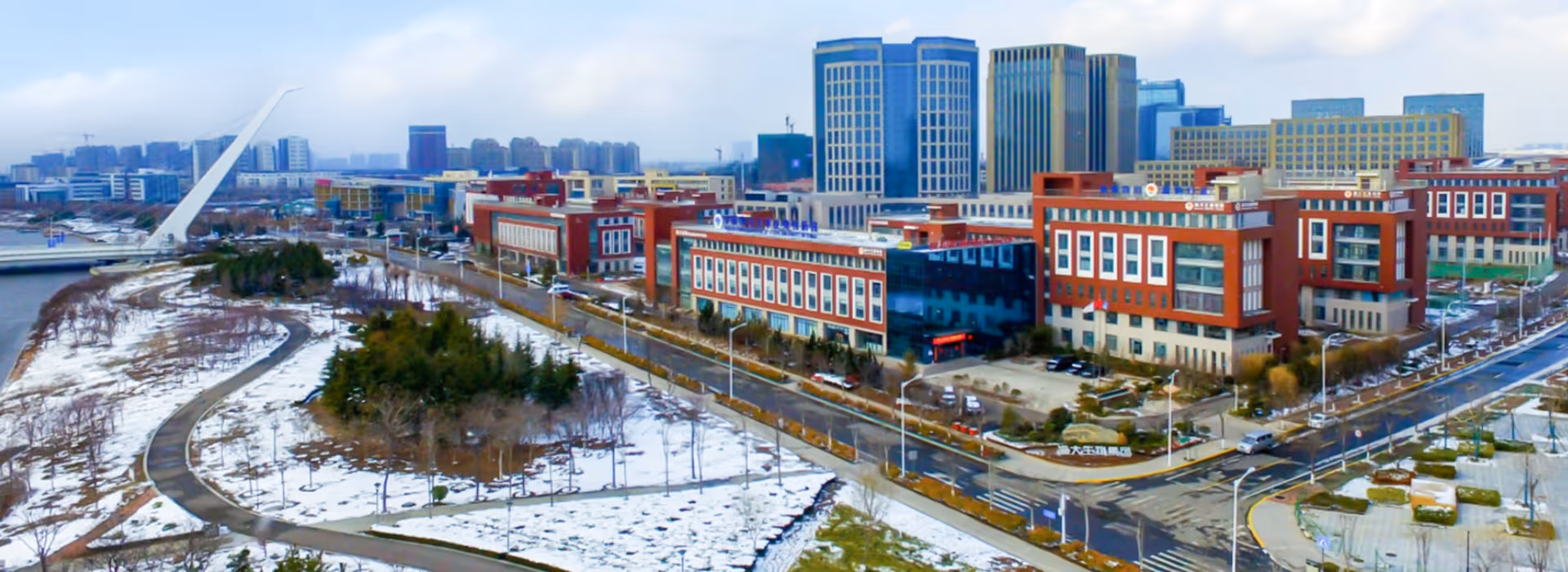 Aerial view of a modern cityscape featuring a distinctive white cable-stayed bridge, snowy park with winding paths, and red brick office buildings among taller skyscrapers.