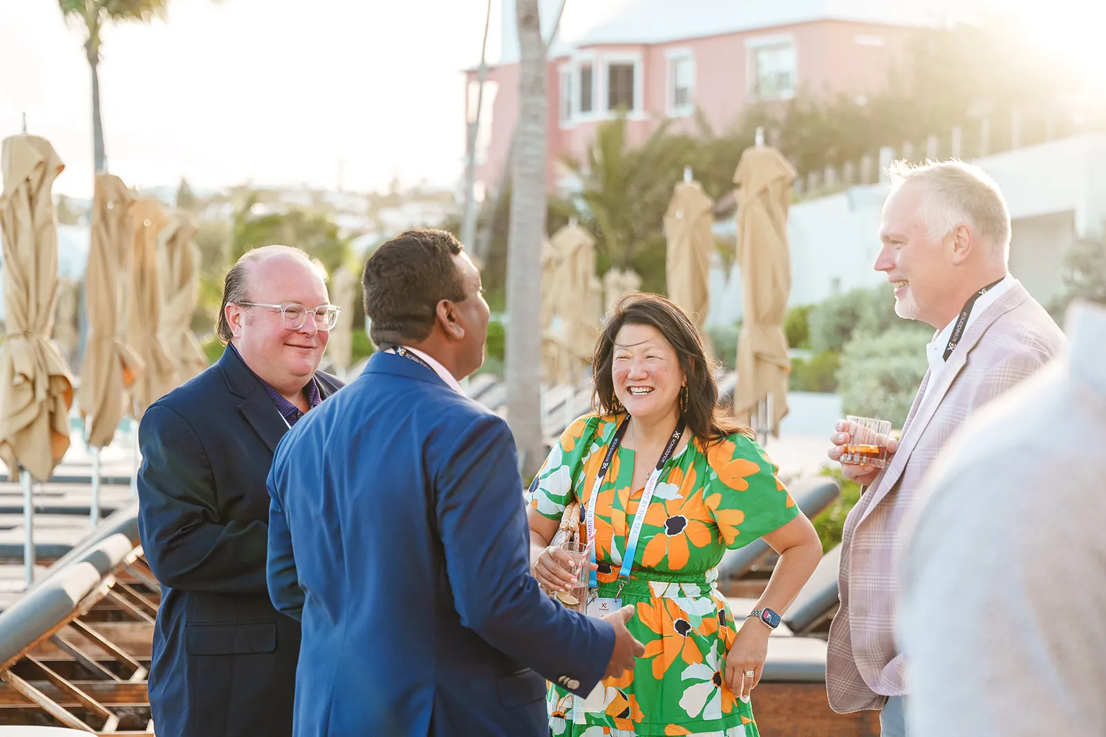 Four people socializing outdoors near lounge chairs and umbrellas, three men and one woman, smiling and holding drinks.