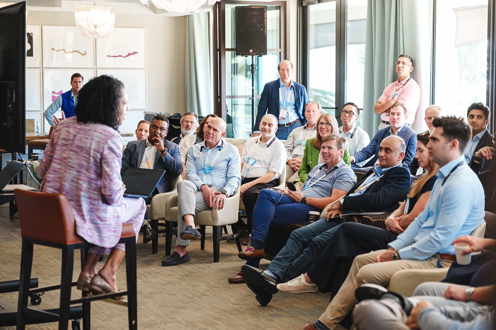 A diverse group of adults attentively listening to a speaker seated on a stool with a laptop in a bright conference room.