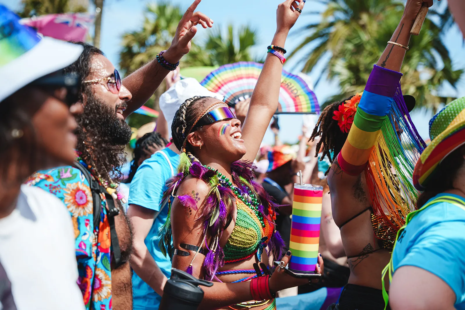 People celebrating at a pride event wearing colorful rainbow attire and accessories under a sunny sky with palm trees.