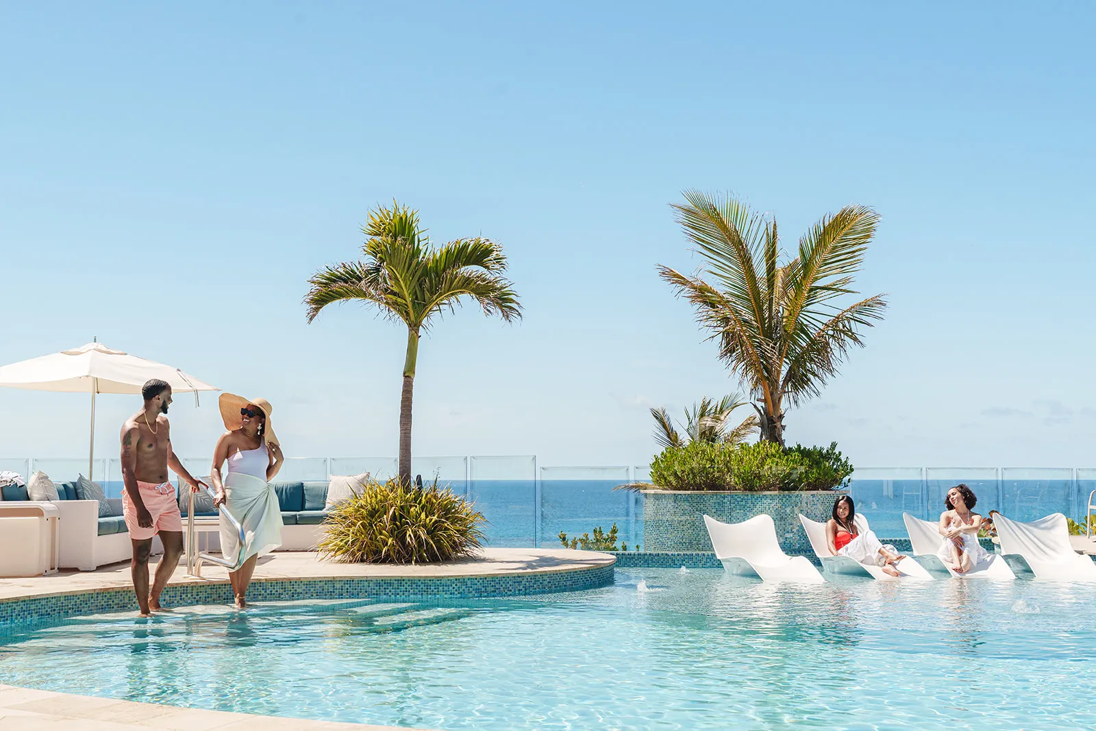 A couple walking in a pool while two women relax on white lounge chairs partially submerged in water with palm trees and ocean in the background.