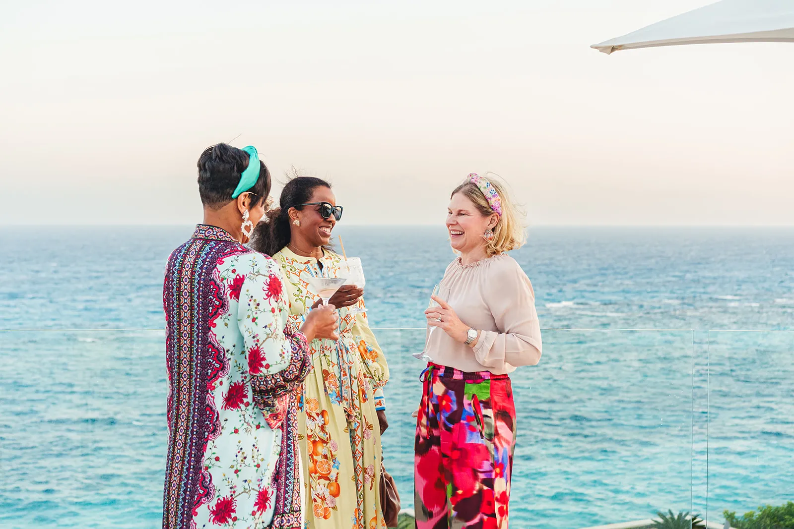 Three women smiling and holding drinks on a terrace overlooking the ocean at sunset.