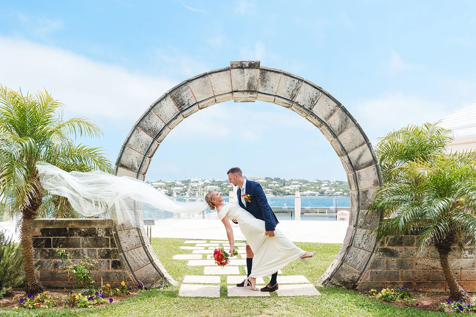 Bride in a white dress with flowing veil and groom in dark suit pose romantically under a stone arch near water.