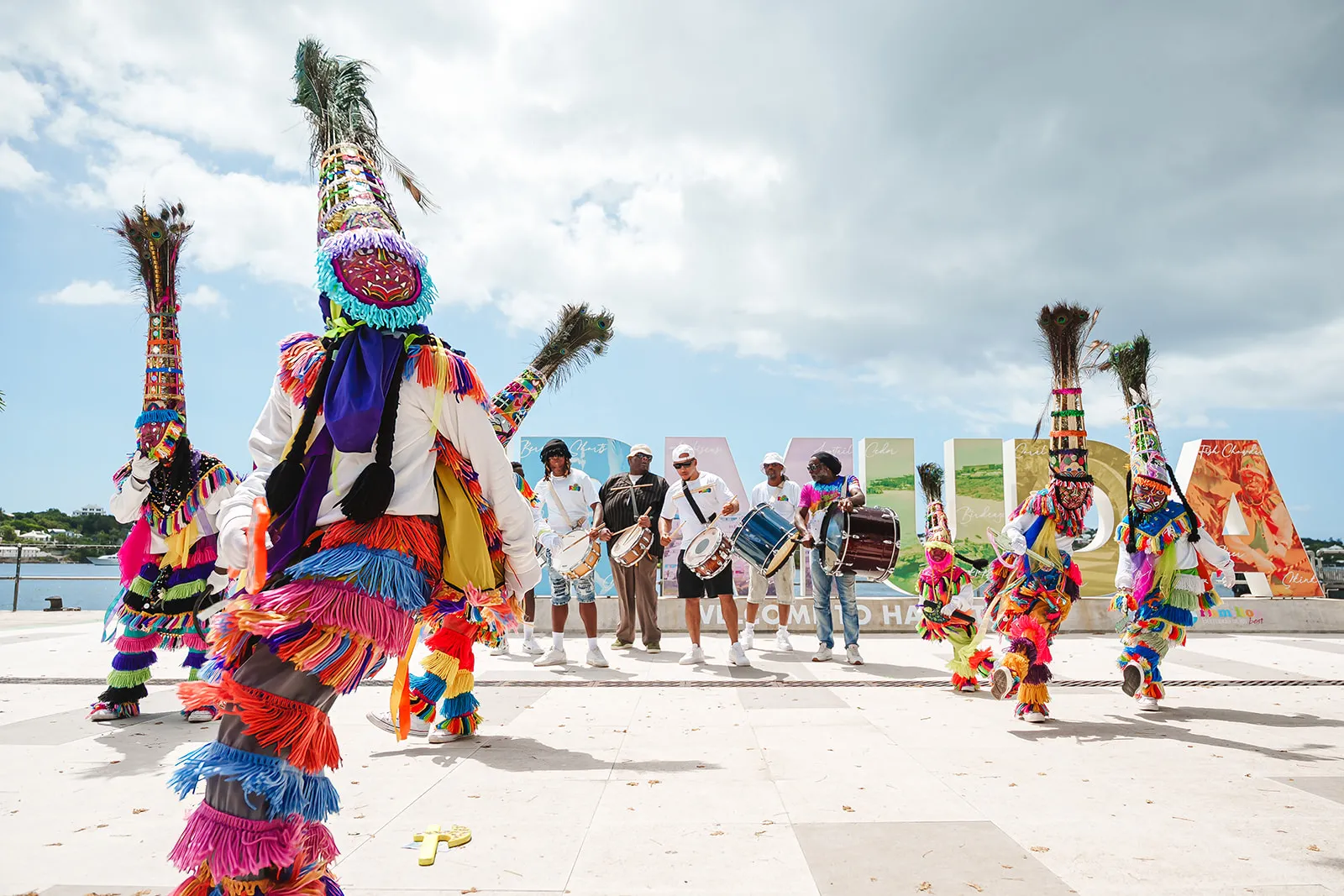 Performers in vibrant, multicolored fringe costumes and tall feathered hats dancing with musicians playing drums in front of large colorful letters spelling 'HALIFAX'.