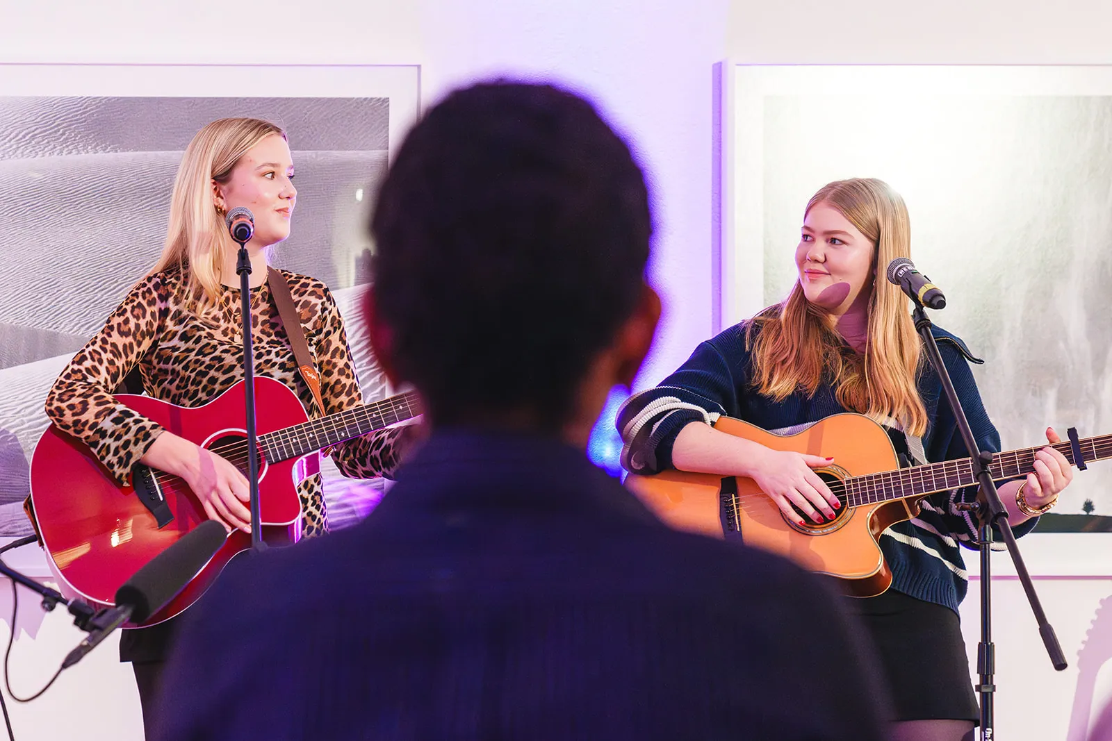 Two young women playing acoustic guitars and singing into microphones during a live indoor performance with an audience member in the foreground.