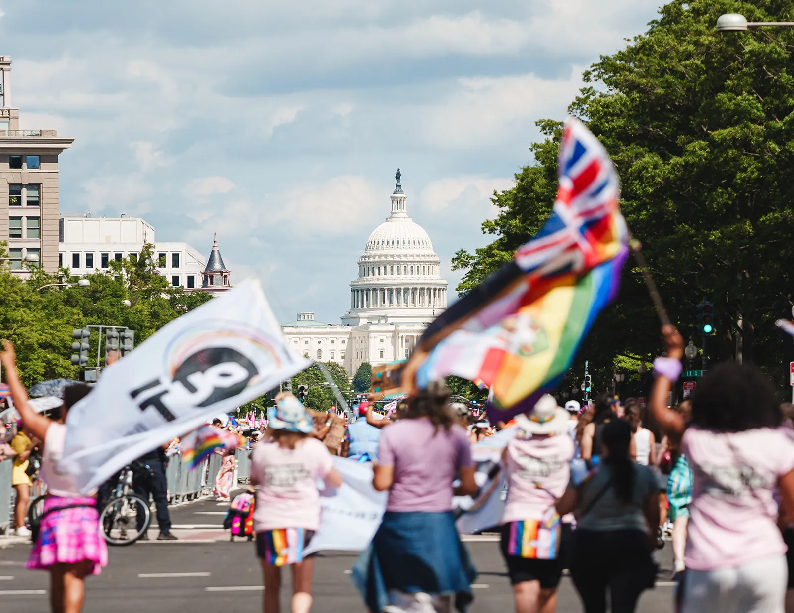 Crowd of people holding rainbow flags and banners walking towards the U.S. Capitol building during a parade or rally.