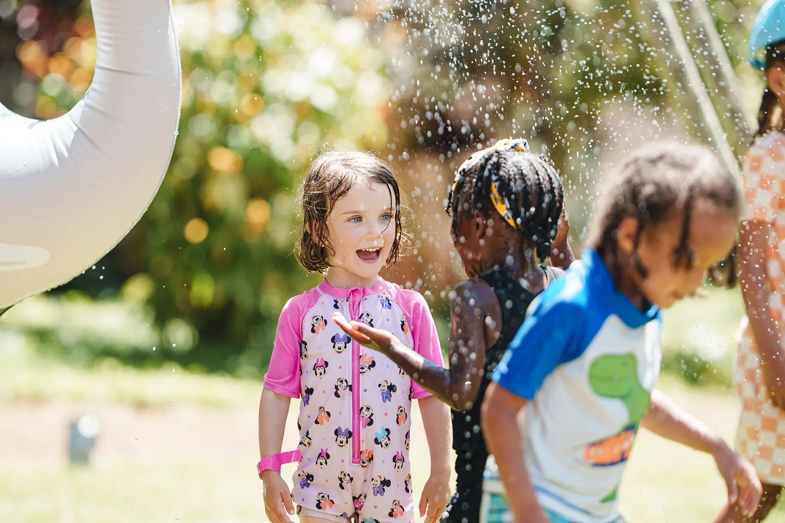 Children playing and laughing outdoors under water spray on a sunny day.