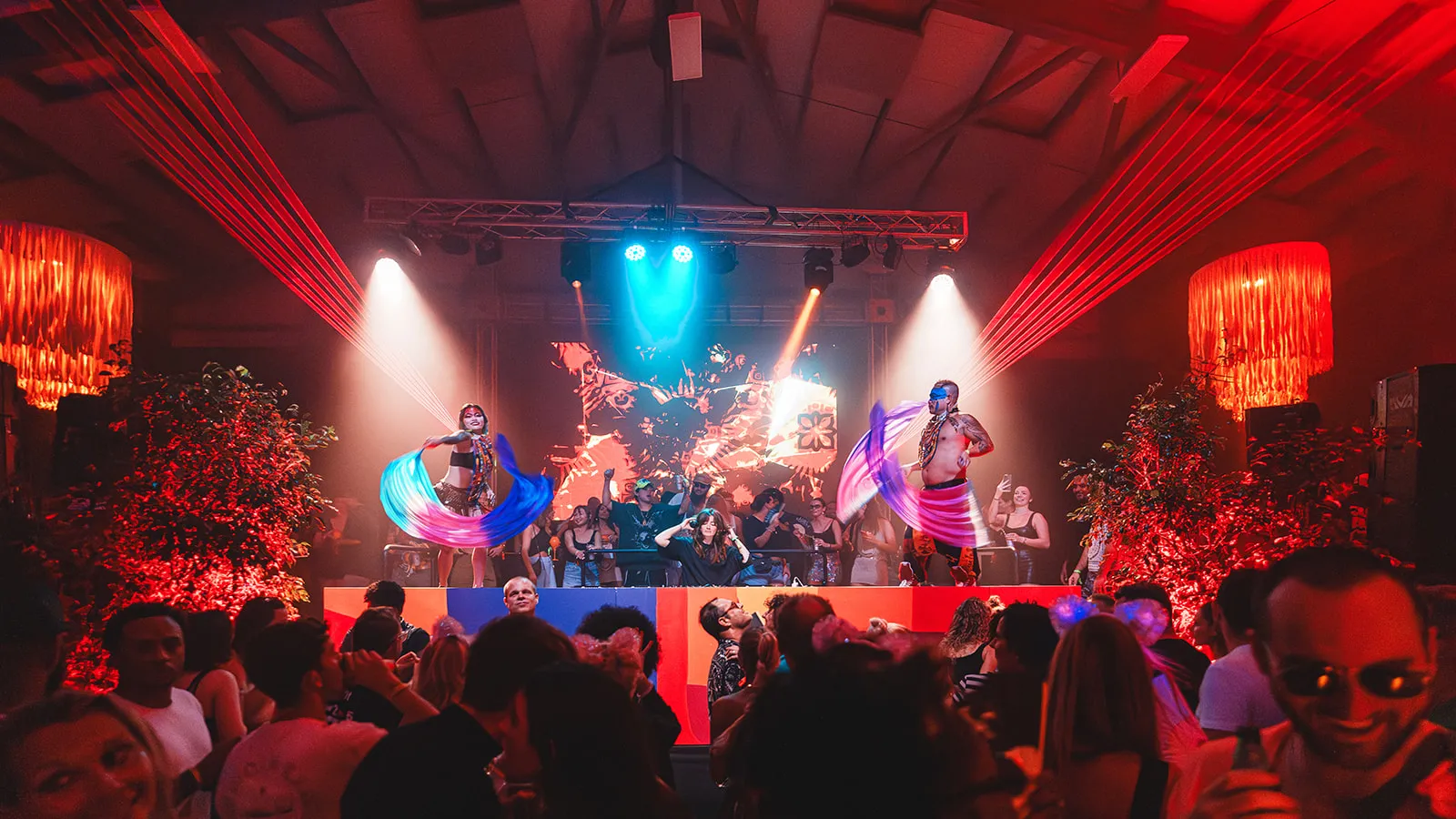 A lively nightclub scene with two performers waving colorful scarves on a stage, a DJ mixing music, and a crowd dancing under red and blue lights.