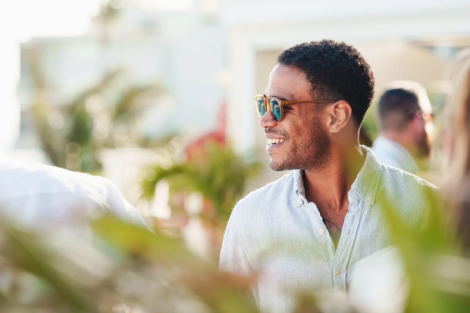 Smiling man wearing sunglasses and a light-colored button-up shirt in an outdoor setting with blurred greenery and people in the background.