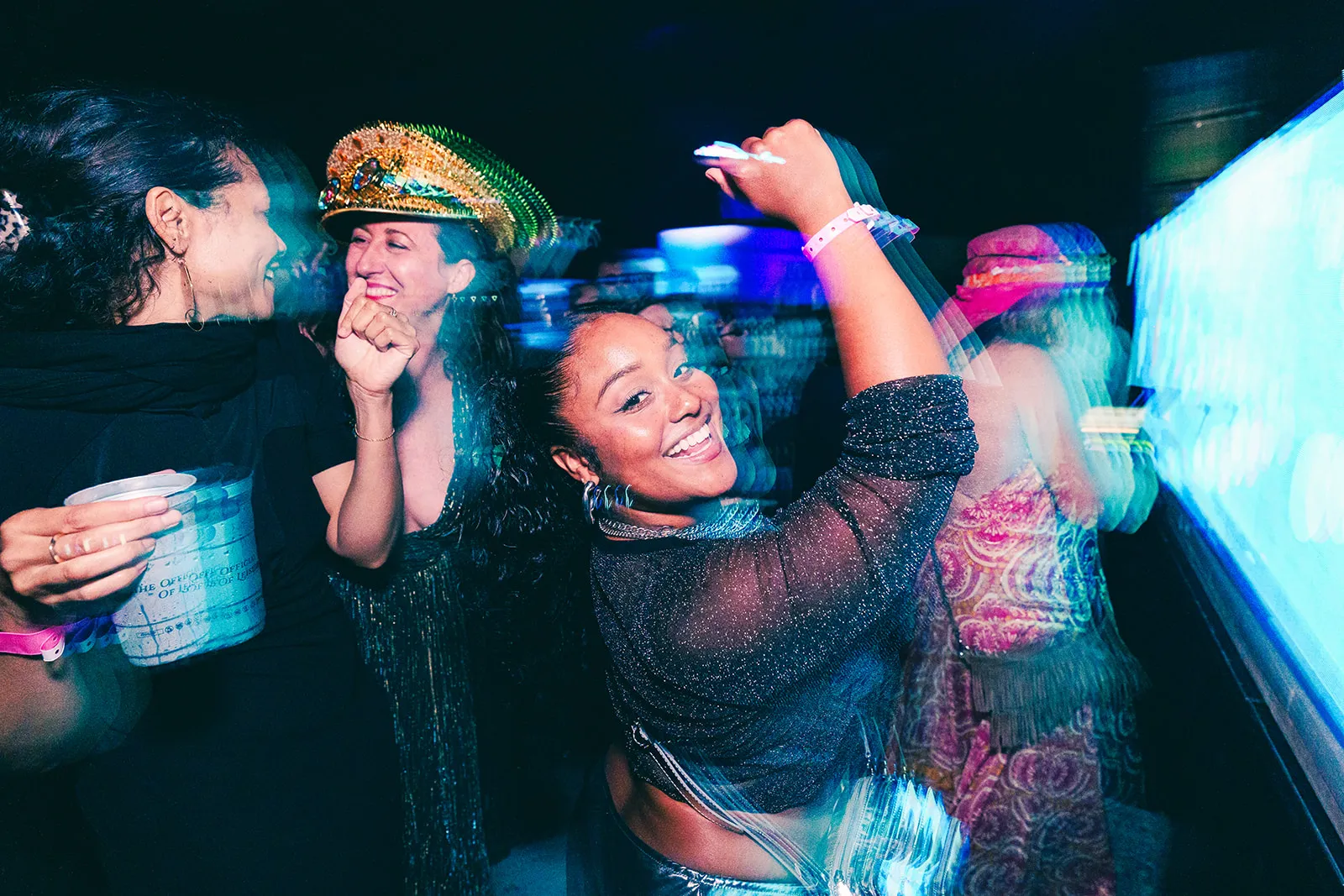 Three women dancing and smiling at a lively party with colorful lighting and festive attire.