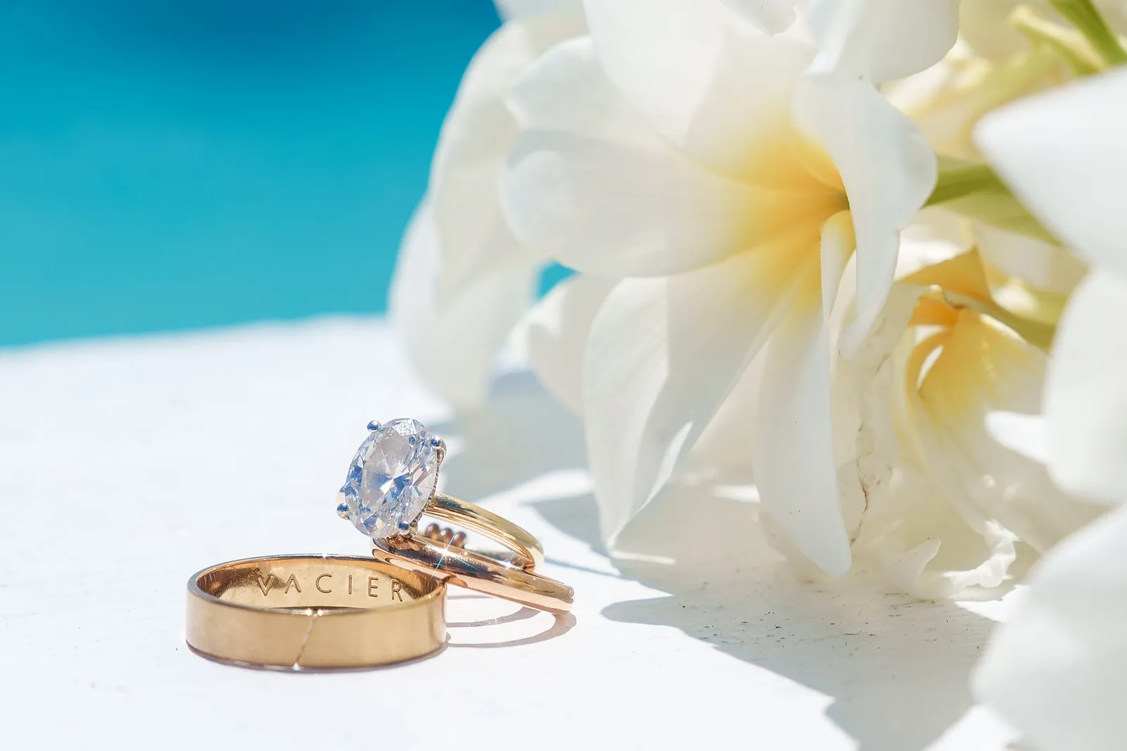 Close-up of three gold rings, one with a large clear gemstone, next to white flowers with yellow centers on a light surface.