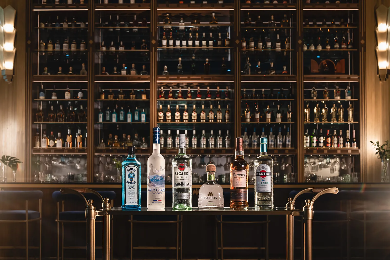 Six bottles of spirits including Bombay Sapphire gin, Grey Goose vodka, Bacardi rum, Patrón silver tequila, Dewar's Scotch whisky, and Martini Bianco vermouth displayed on a bar cart in front of a stocked bar.