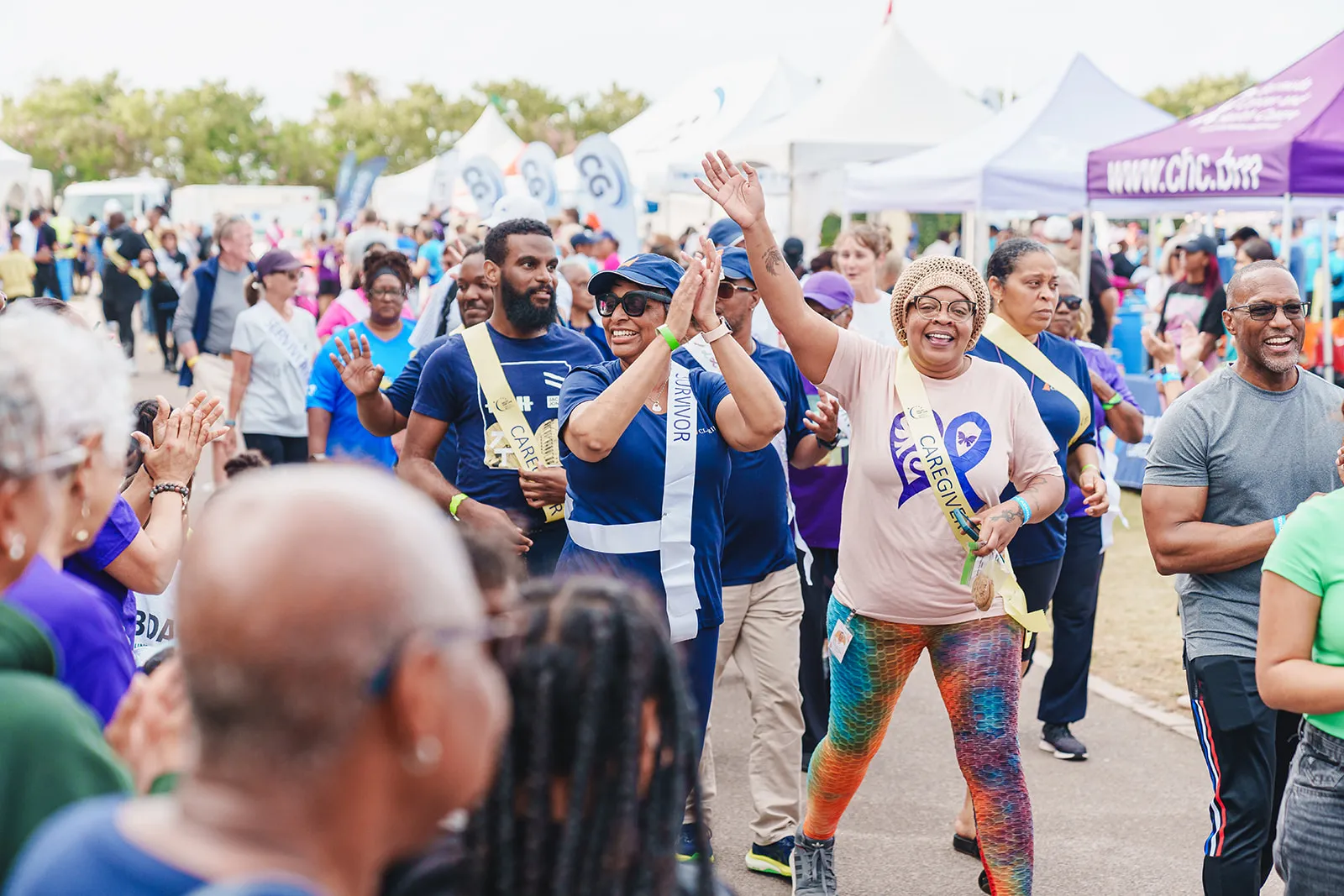 People walking and celebrating outdoors at a Relay for Life event with some wearing survivor and caregiver sashes.