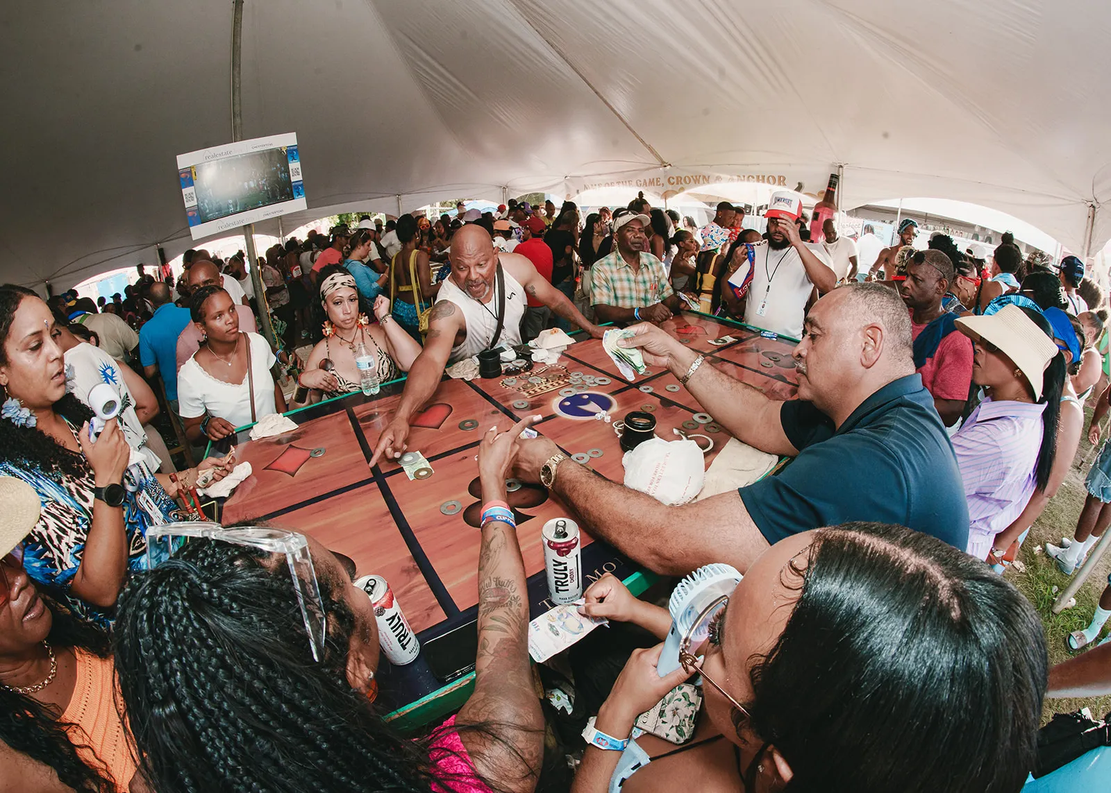 People playing a competitive game involving sliding coins on a wooden board under a large tent during a crowded outdoor event.