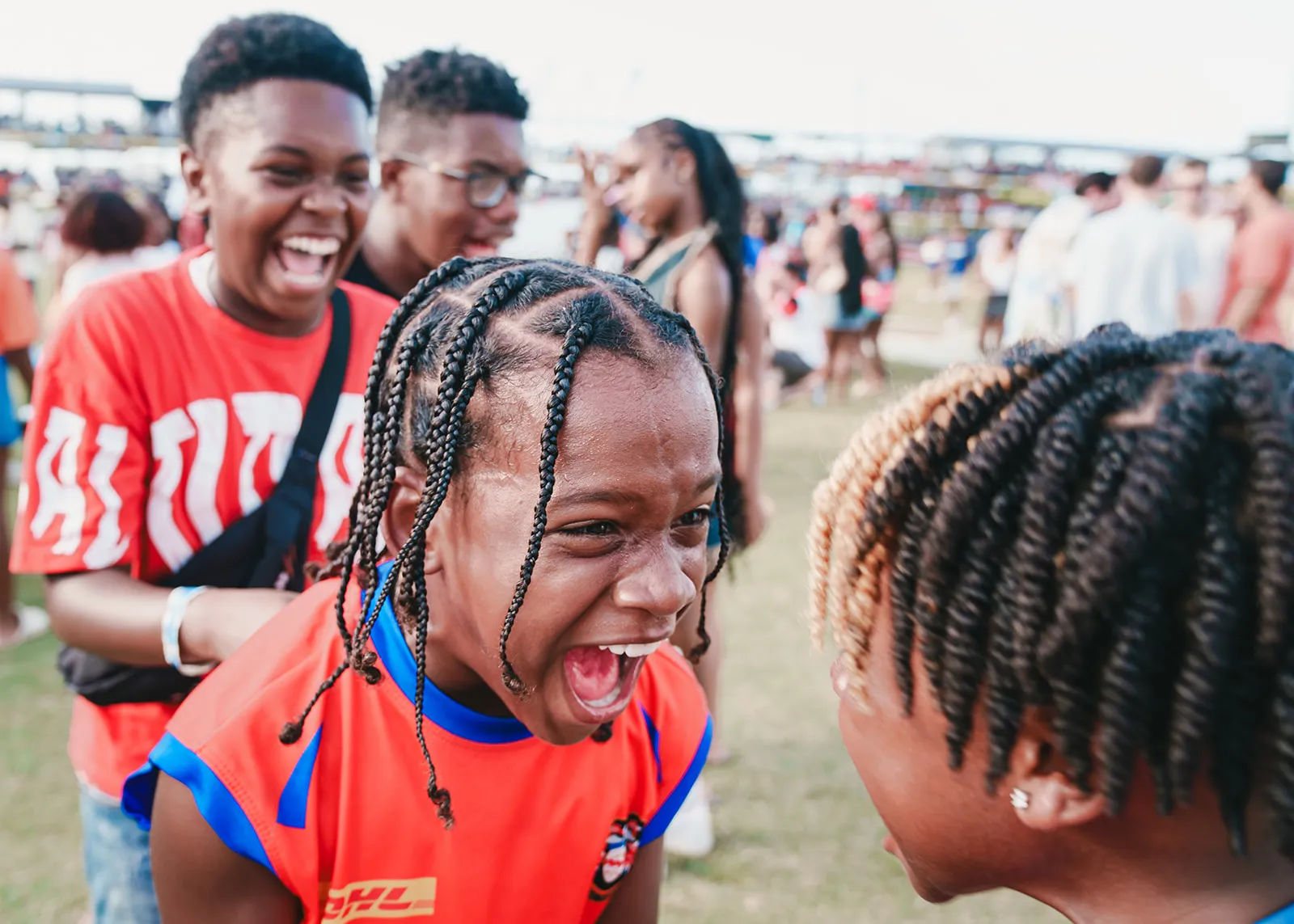 Children with braided hair excitedly interacting at an outdoor event with a crowd in the background.