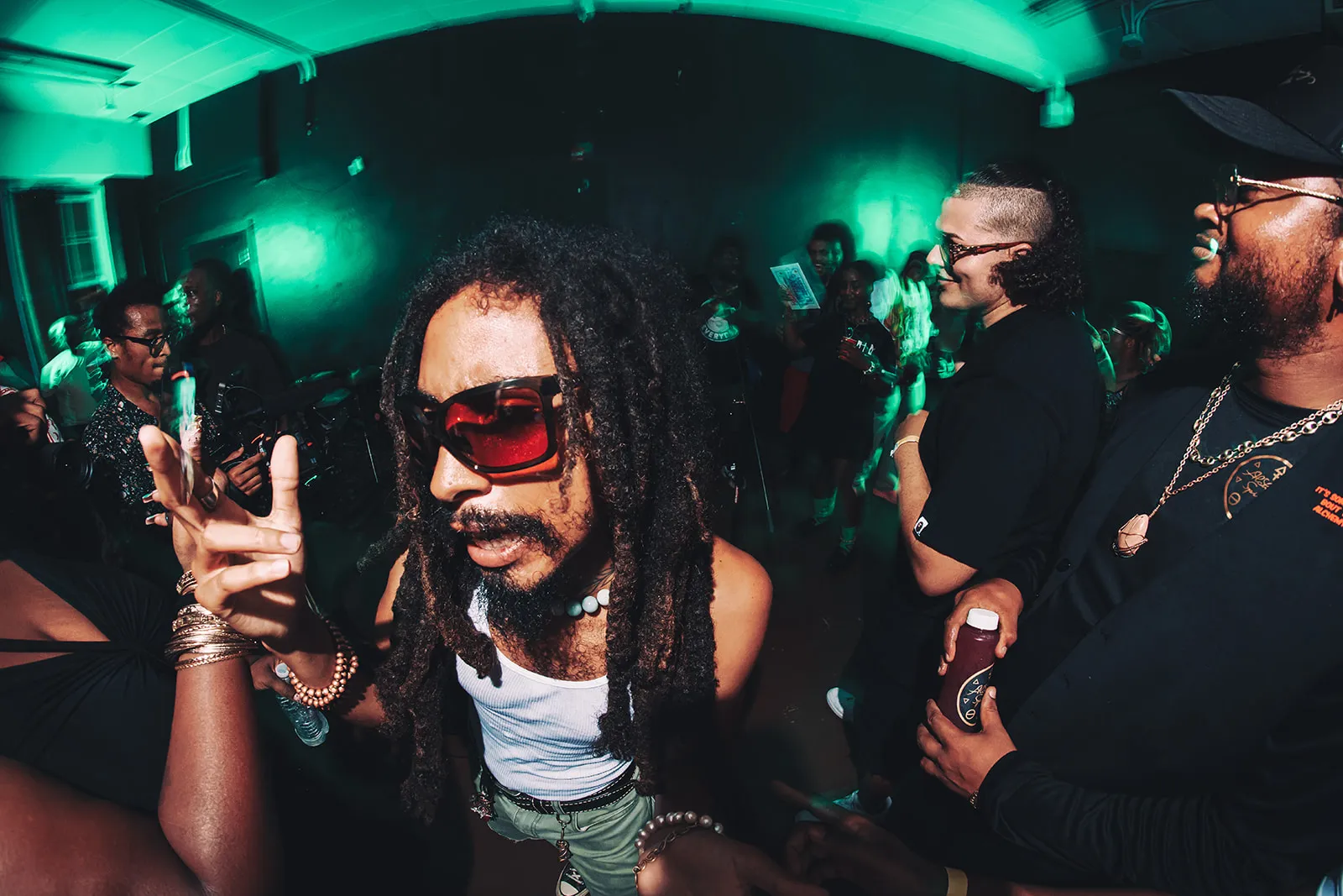 Person with dreadlocks and red sunglasses dancing at a dimly-lit party with other people in the background.