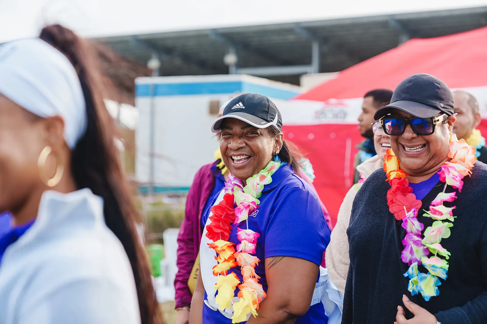 Two women smiling and wearing colorful leis at an outdoor event with a red tent in the background.