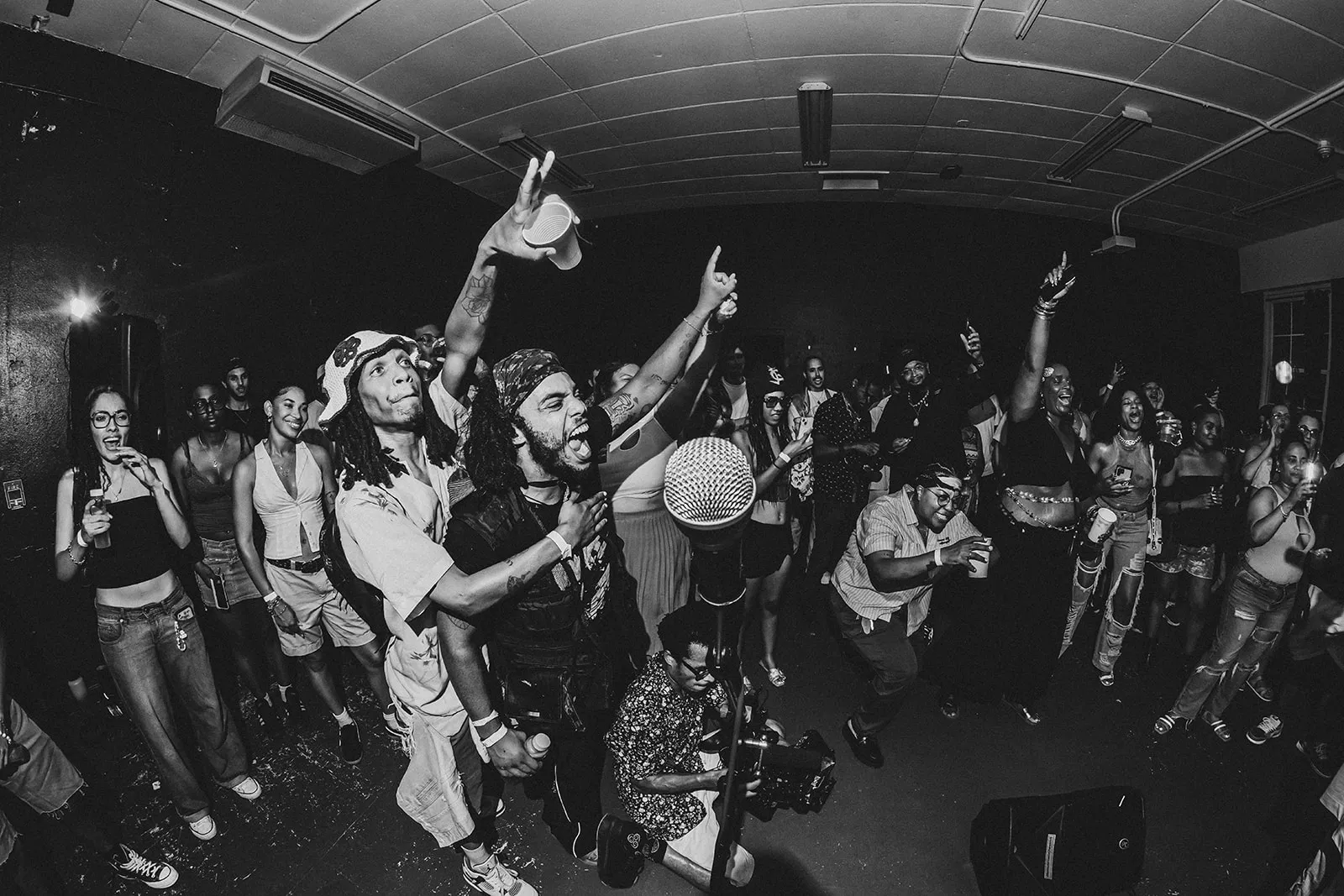Energetic crowd enjoying a lively indoor music event with people singing and dancing around a microphone.