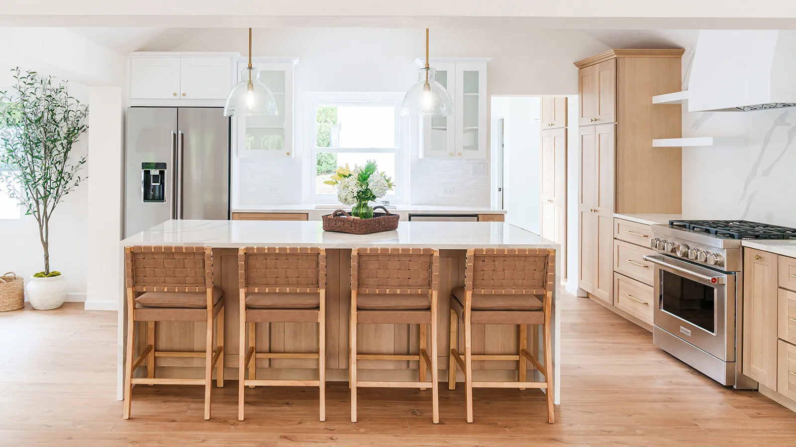Bright modern kitchen with white countertops, wooden cabinetry, four woven-back bar stools, stainless steel refrigerator and stove, and a green plant in a white pot.