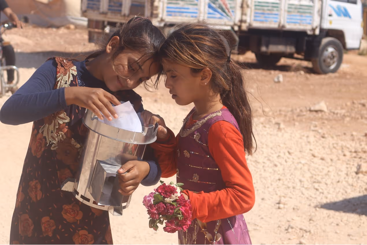 Two young girls, one holding a bouquet of pink flowers, putting a piece of paper into a metal collection box outdoors.