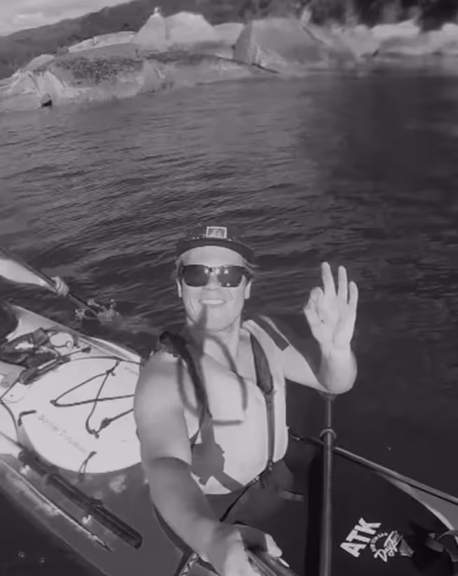 Smiling man wearing sunglasses and a cap, kayaking on calm water near rocky shores, waving at the camera.