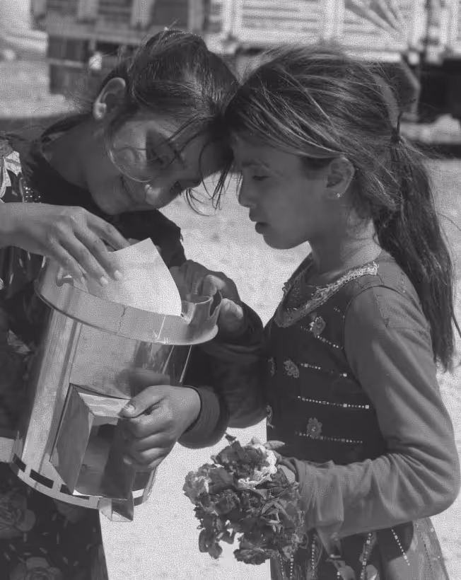 Two young girls, one holding flowers, looking inside a metal container together.
