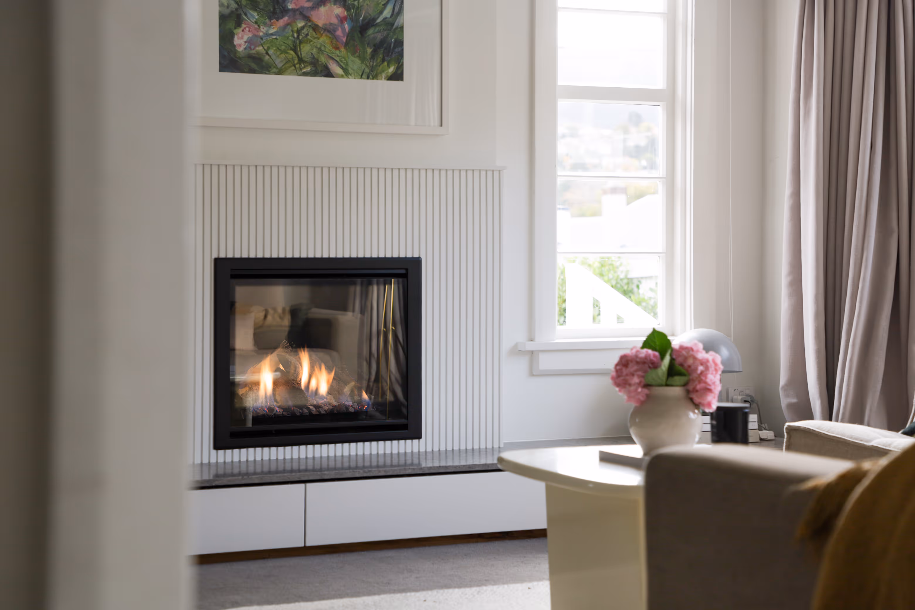 Modern gas fireplace with realistic burning logs set in a light-coloured wall next to a wooden side table with books.