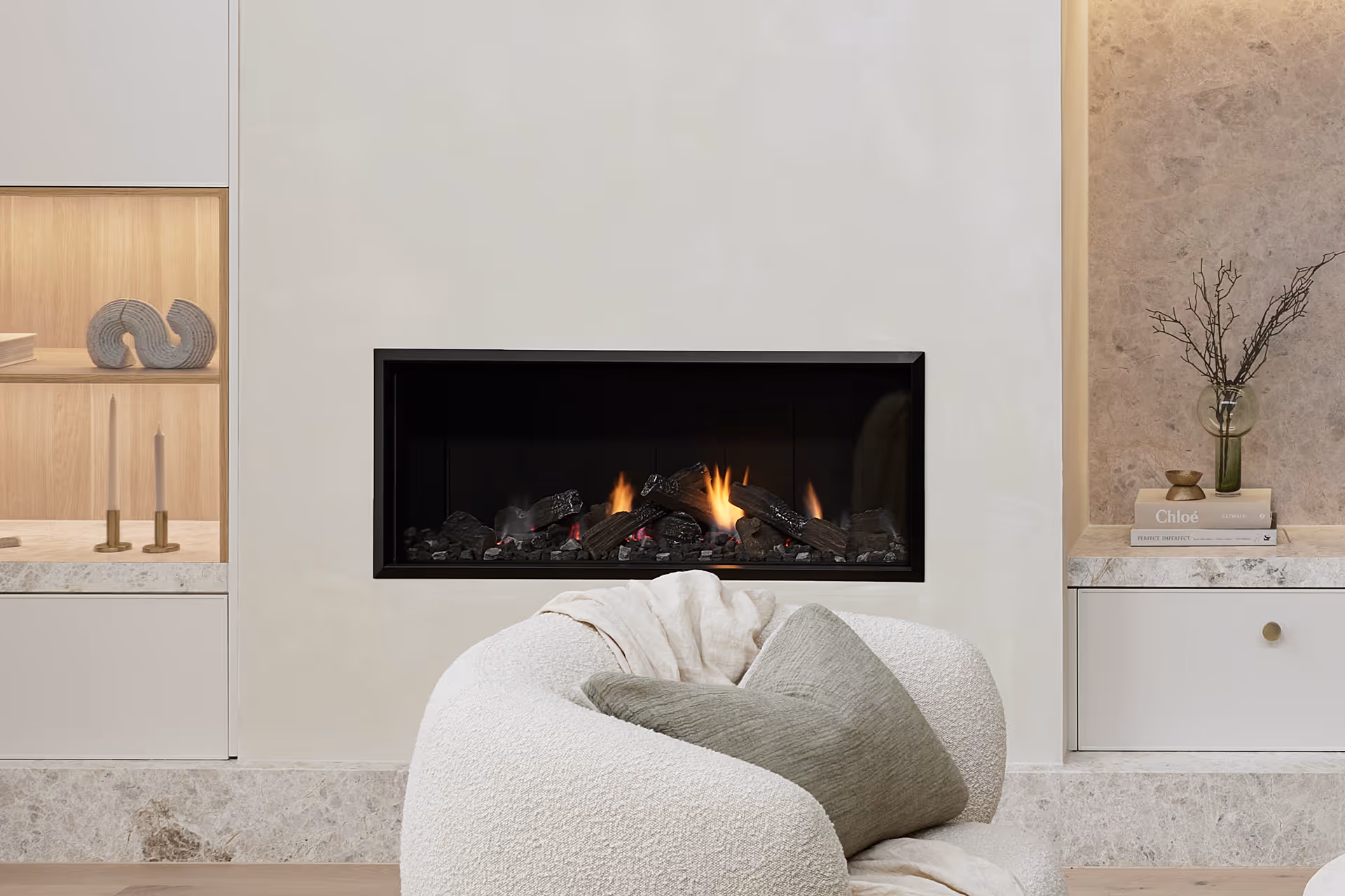 Cheminée à gaz moderne avec des bûches réalistes, encastrée dans un mur de couleur claire, à côté d'une table d'appoint en bois avec des livres.