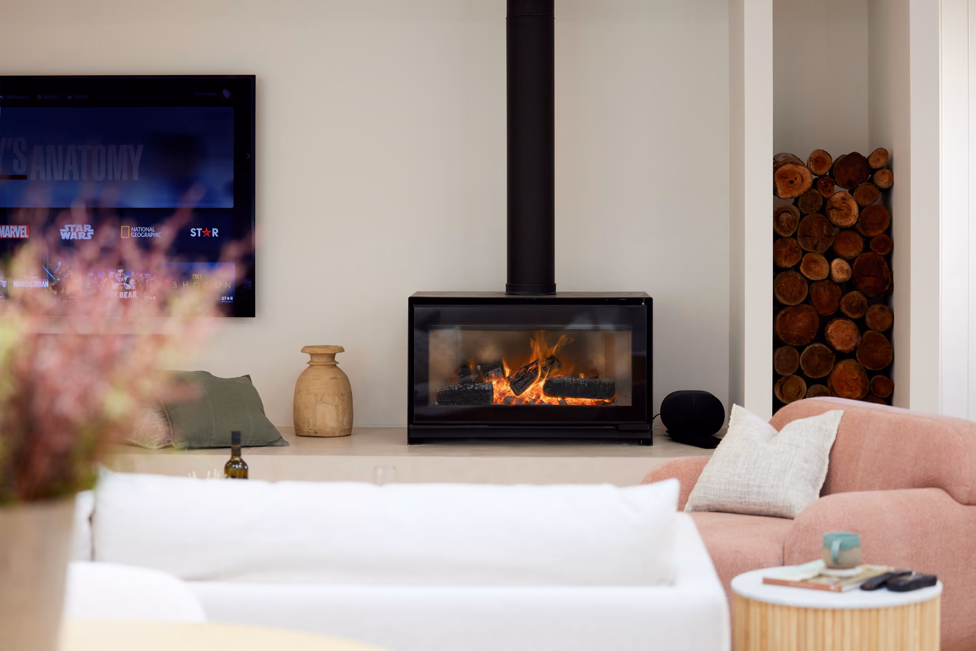 Modern gas fireplace with realistic burning logs set in a light-coloured wall next to a wooden side table with books.
