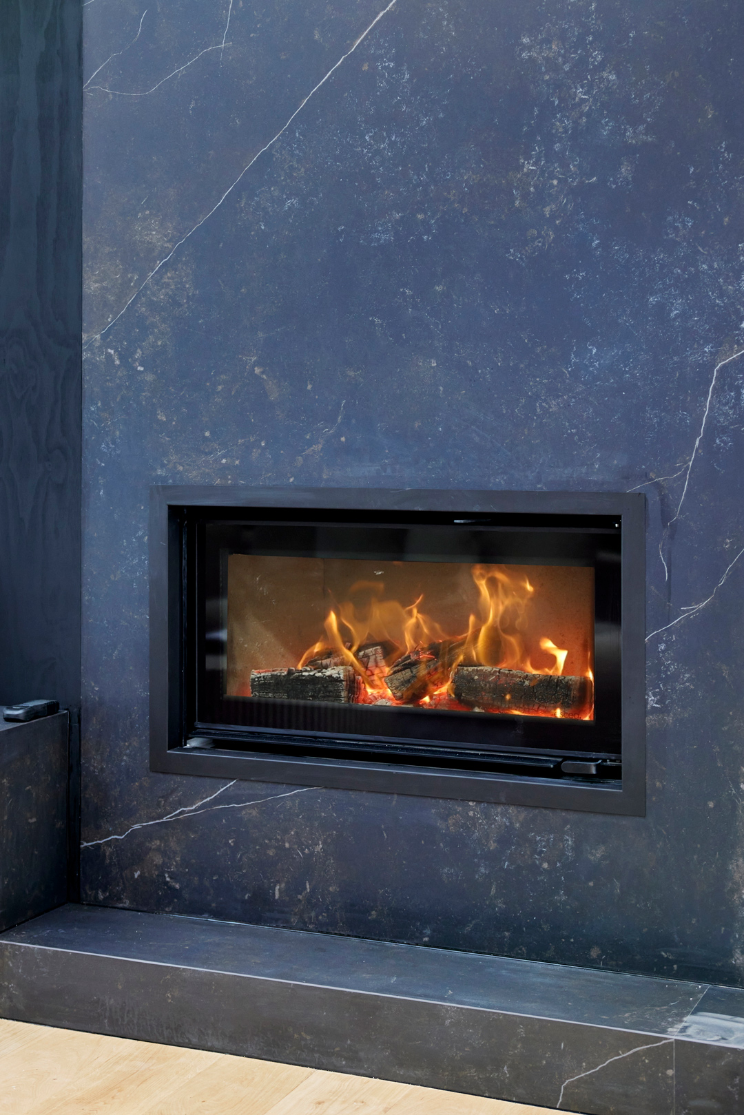 Modern gas fireplace with realistic burning logs set in a light-coloured wall next to a wooden side table with books.