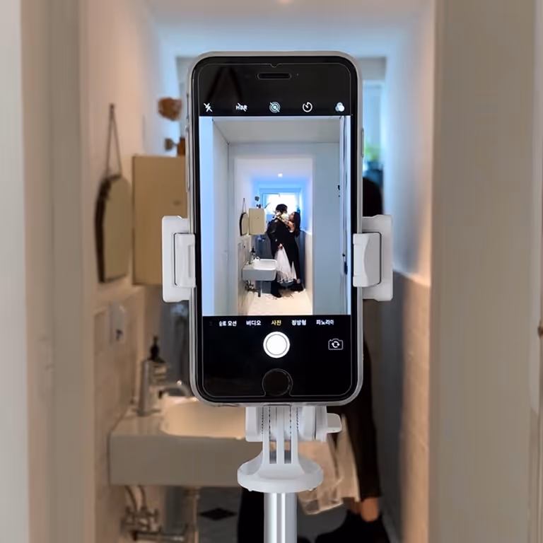 Un smartphone fixé sur un trépied prenant en photo un couple s'embrassant dans un petit couloir lumineux avec lavabo.