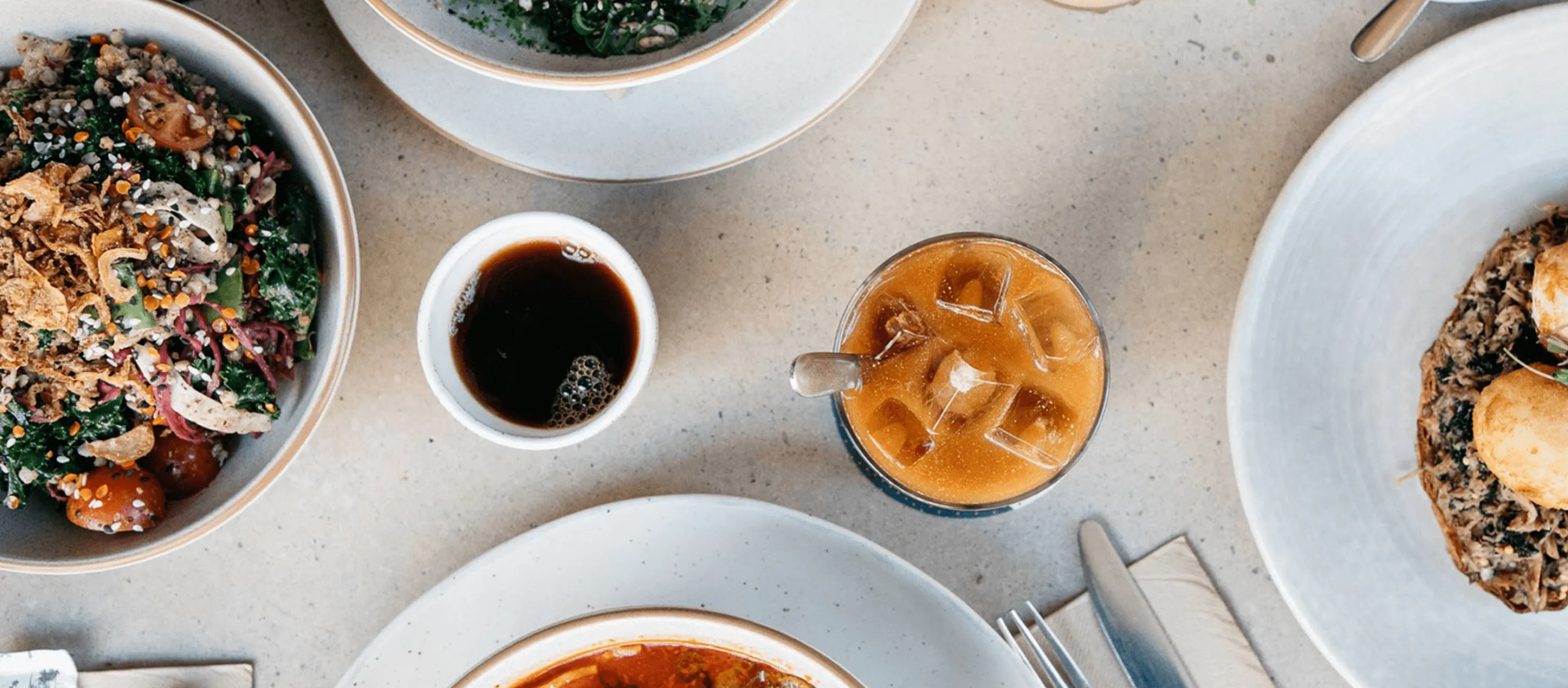 Table set with a bowl of leafy salad, an iced coffee, a cup of black coffee, and plates with food on a light surface.