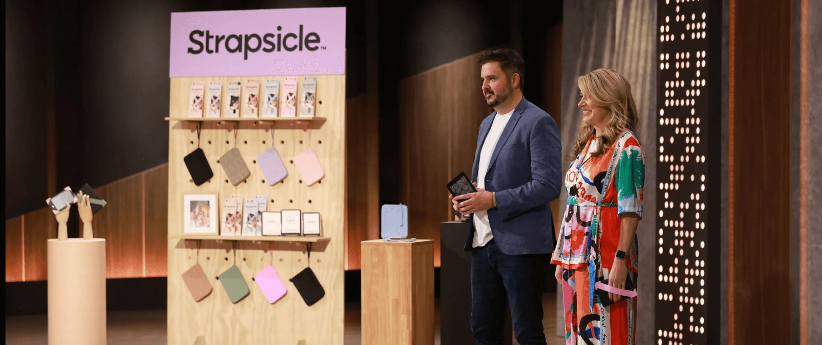 Two presenters stand by a display of colorful phone cases labeled Strapsicle in a studio setting.