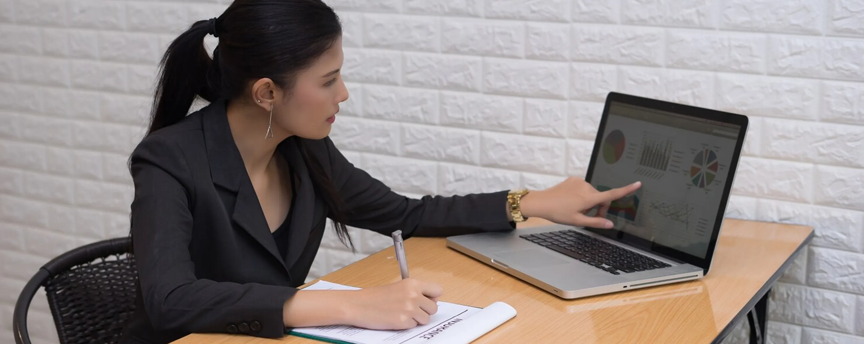 Woman in black blazer pointing at graphs on laptop screen while taking notes at wooden desk.