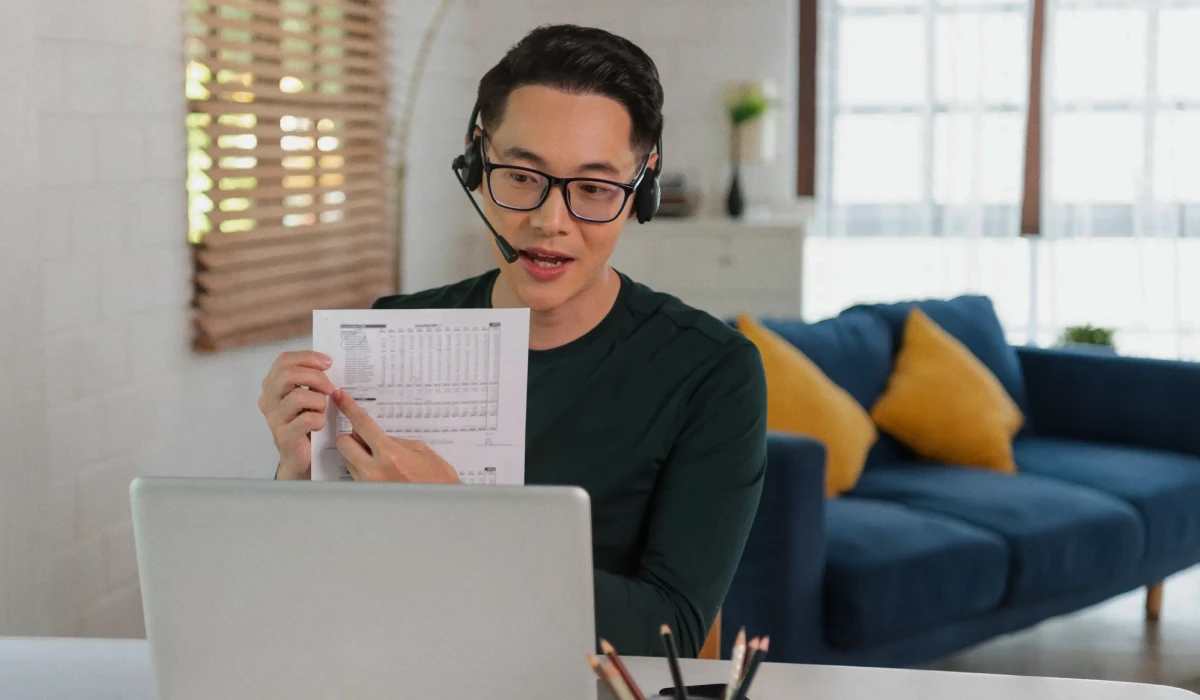 Man wearing glasses and headset pointing at a document while speaking in front of a laptop in a living room.
